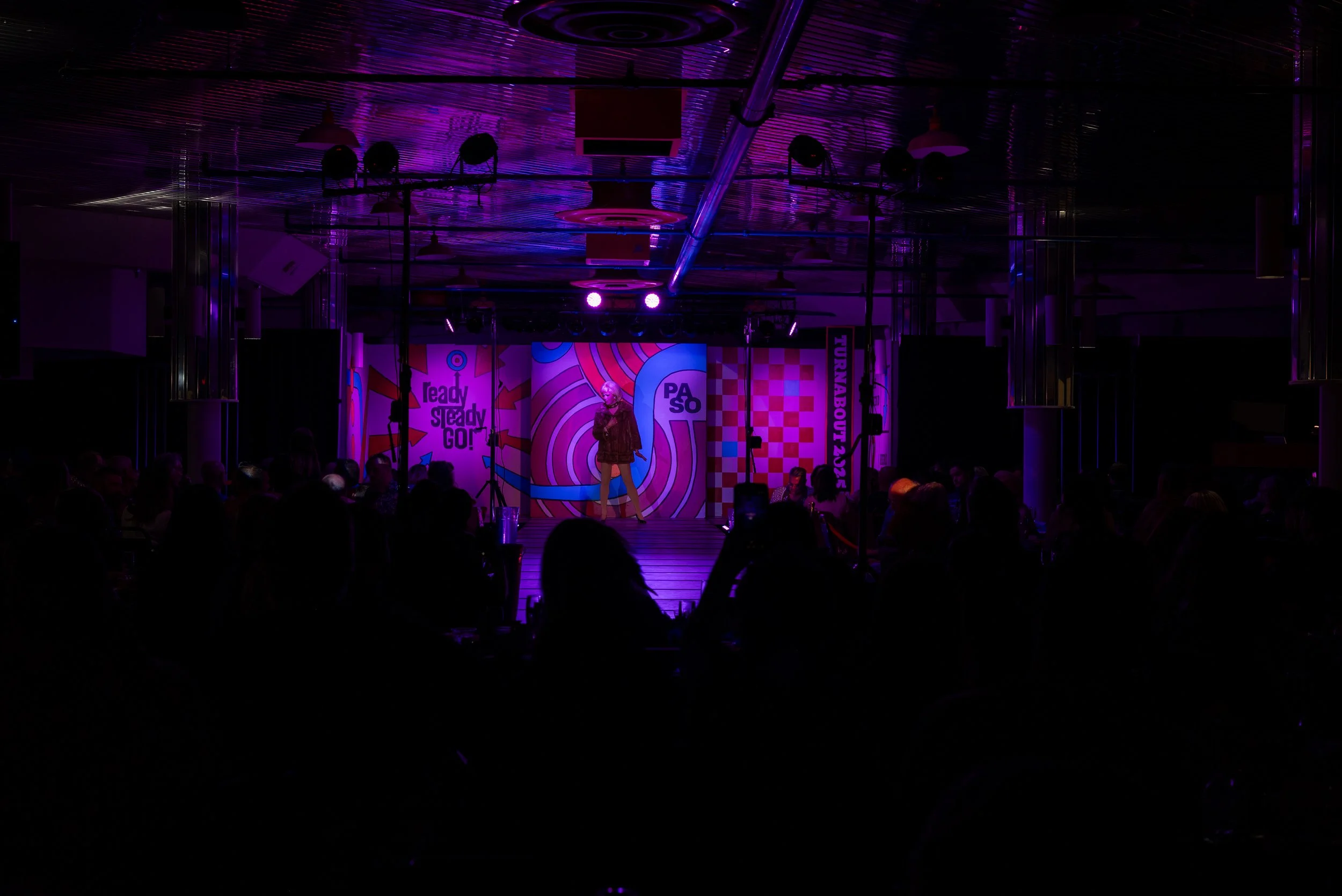 Stage with comedian performing under purple and pink lights, audience seated in front, colorful backdrop with patterns and the text 'ready Steady GO!' and 'PASO'.