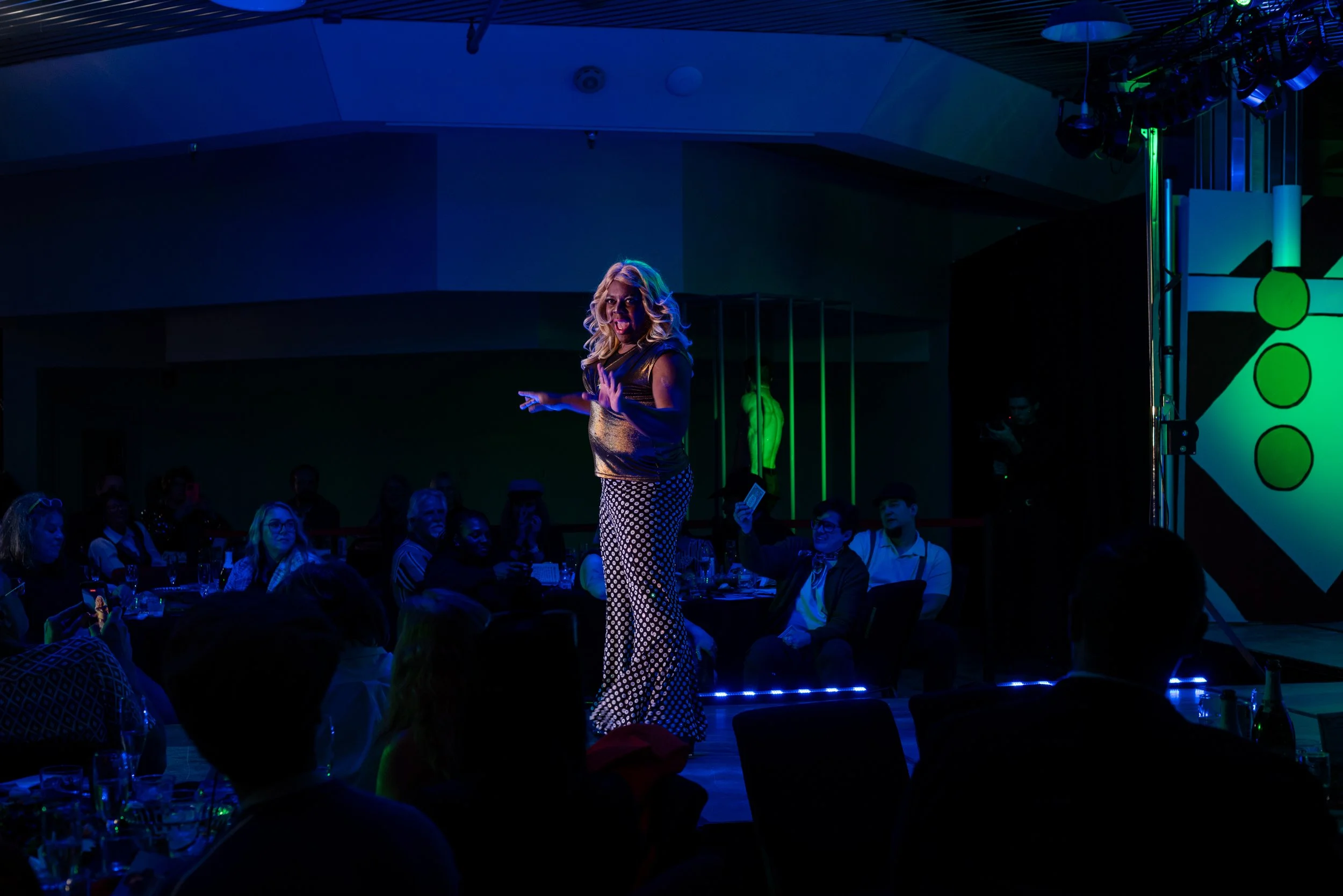 A woman performs on stage at a dark, colorful event, with an audience watching and some taking photos.