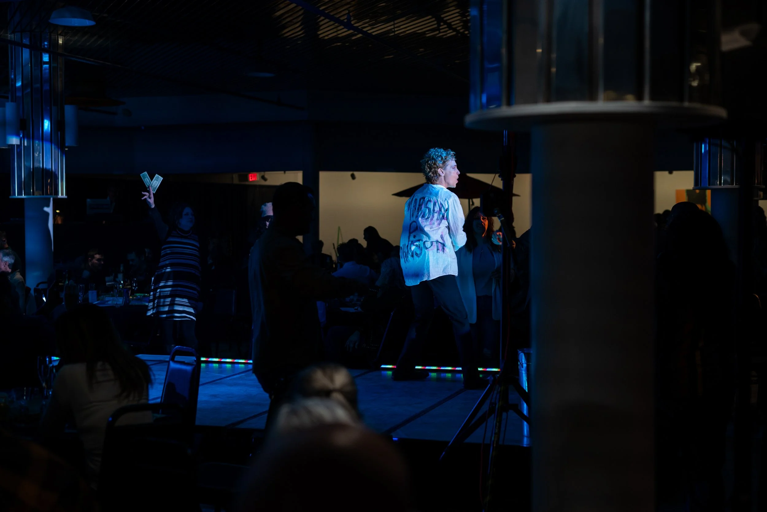 A performer with curly hair standing on stage illuminated by blue lights at an indoor event, with audience members seated and others standing around in a darkened room.