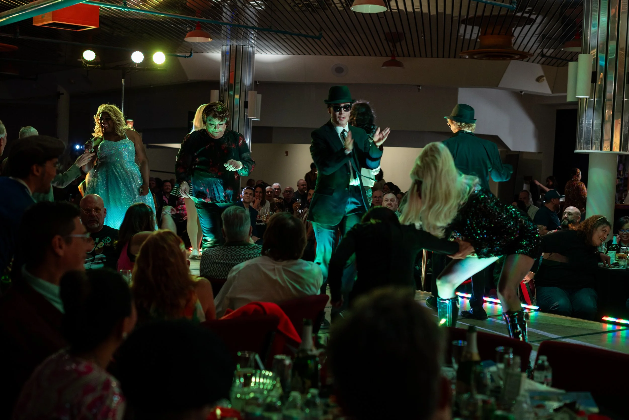 People dancing and singing on stage in a dimly lit venue with an audience seated at tables.