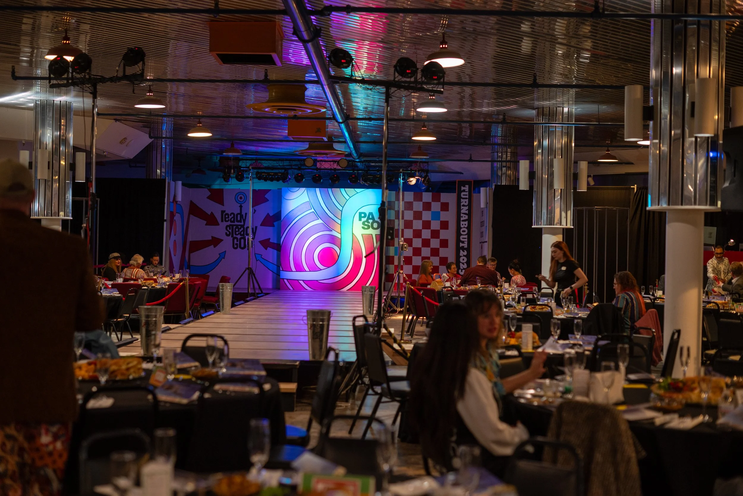 An indoor event with round tables set for dining, a stage with colorful abstract artwork and lighting, and people seated and walking around.