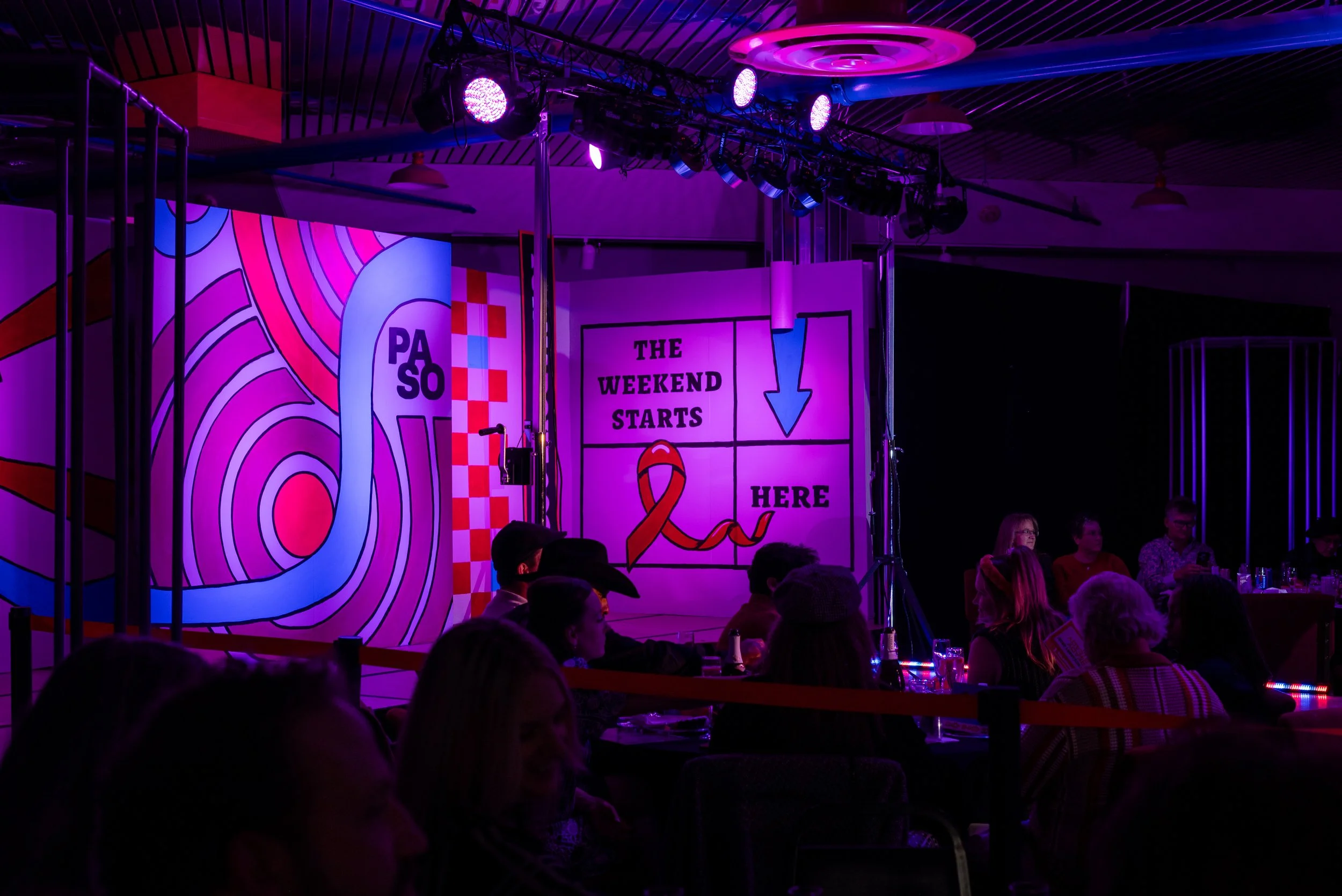 Stage with colorful backdrop and illuminated signs. One sign reads "The weekend starts here" and features a red ribbon, indicating a health-related campaign. The audience is seated in a dark room with purple and pink lighting.