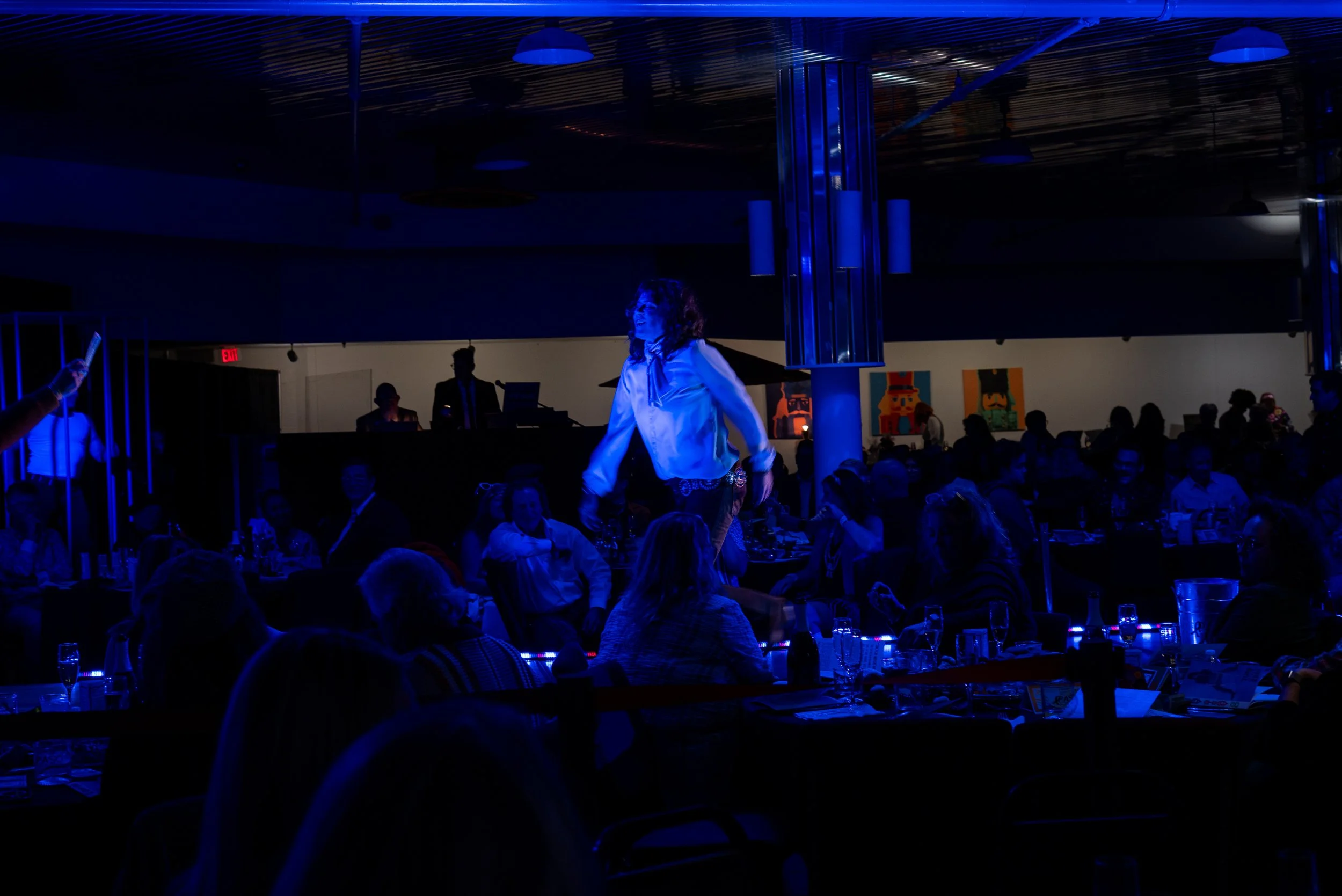 A performer on stage in a dimly lit indoor venue, illuminated by blue lighting, surrounded by an audience seated at tables.