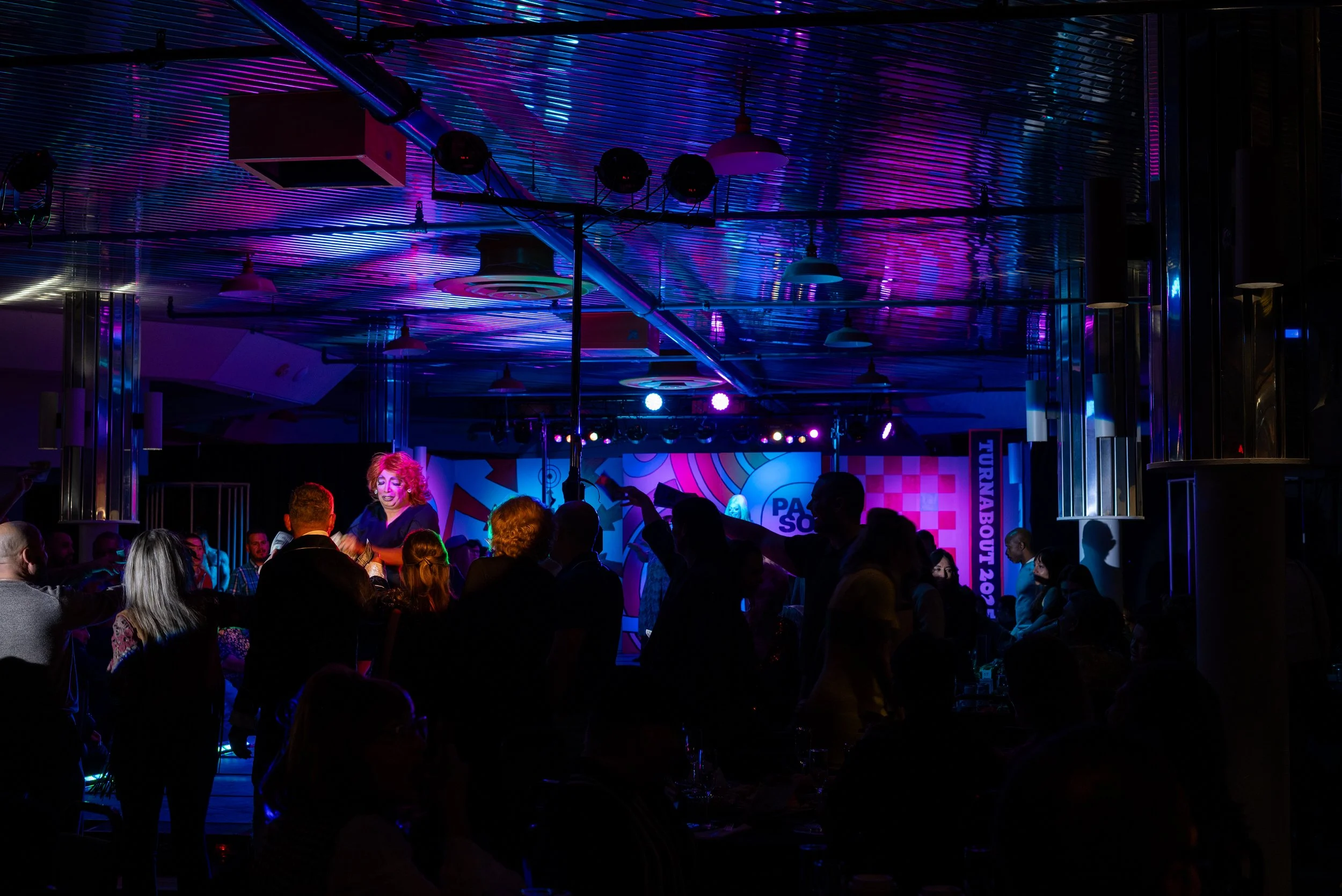 People dancing and socializing at a nightclub or concert with vibrant purple and blue lighting, a stage with a performer, and a colorful background.