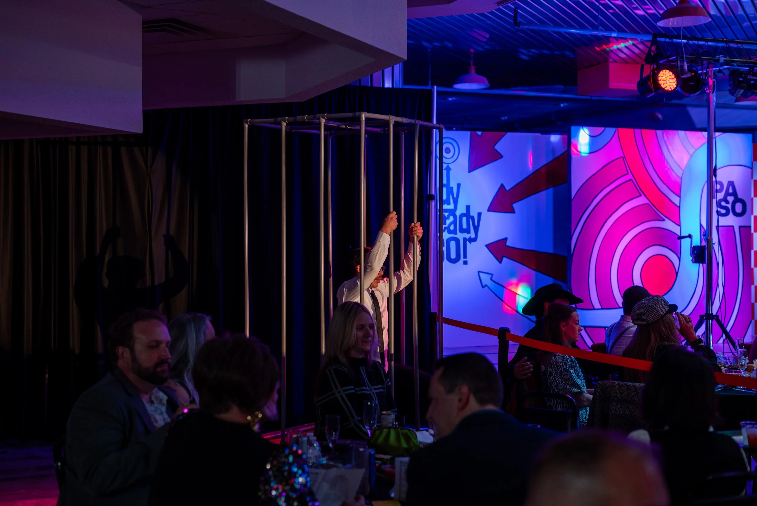 A person in a white shirt and black tie stands inside metallic bars on a stage at a lively event with colorful lighting and a large graphic backdrop.