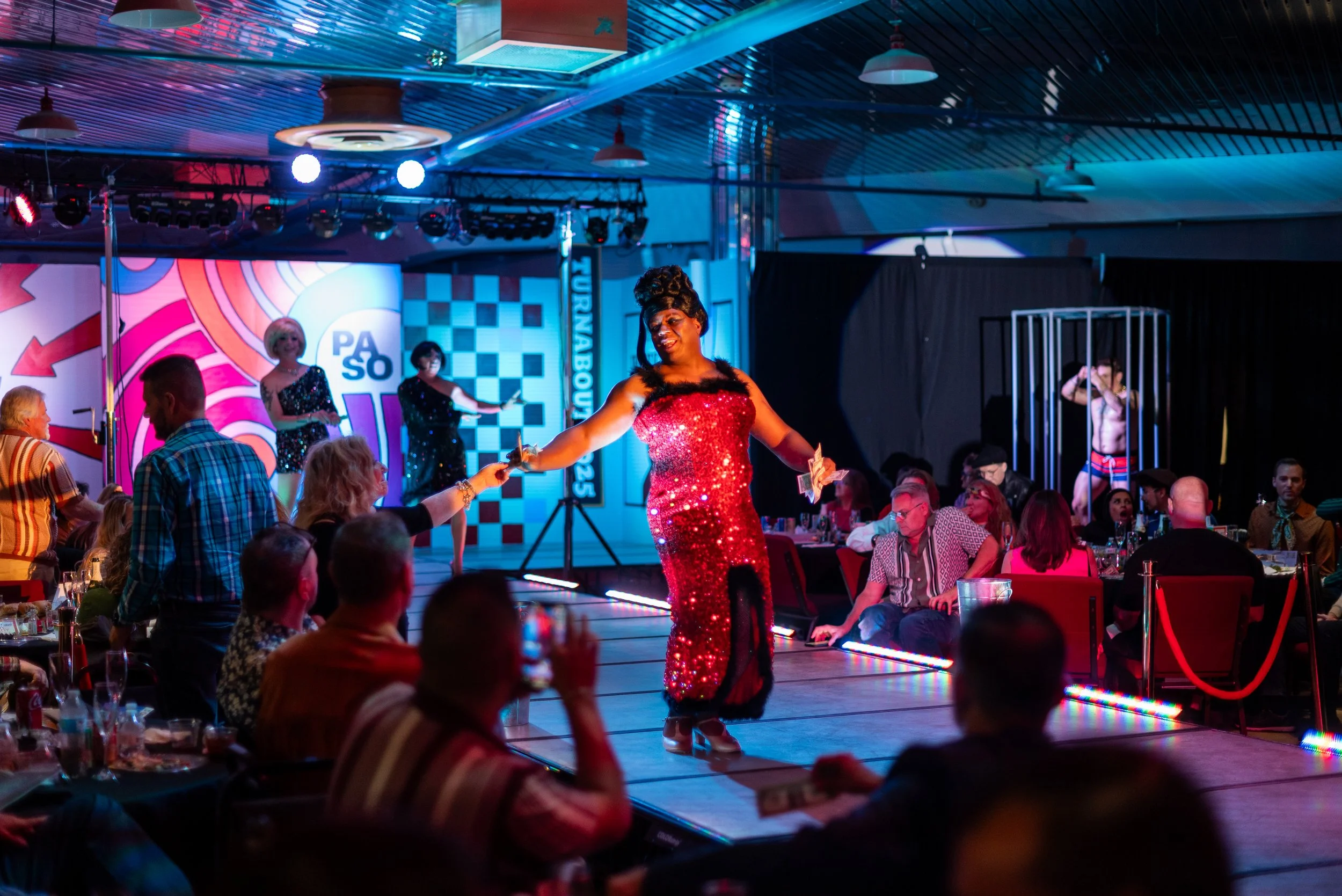 A drag performer in a red sequined dress performing on a stage at a lively event with a colorful backdrop and an audience seated at tables.