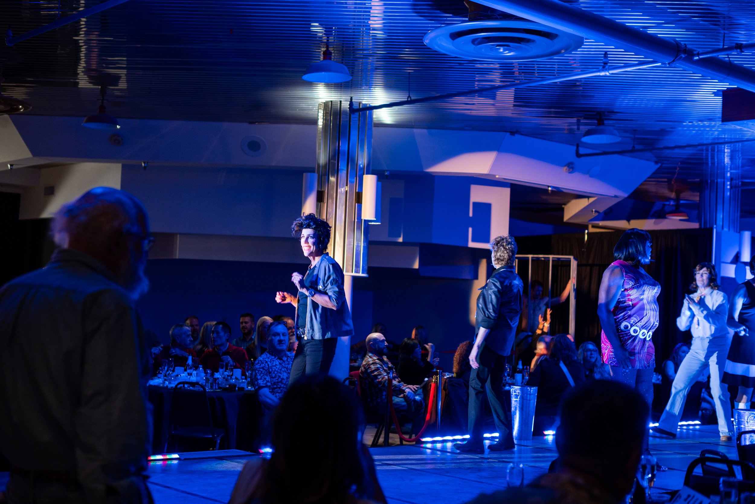 People performing on a dimly lit stage at a show or event, with one woman center stage smiling and others around her, audience seated in front.