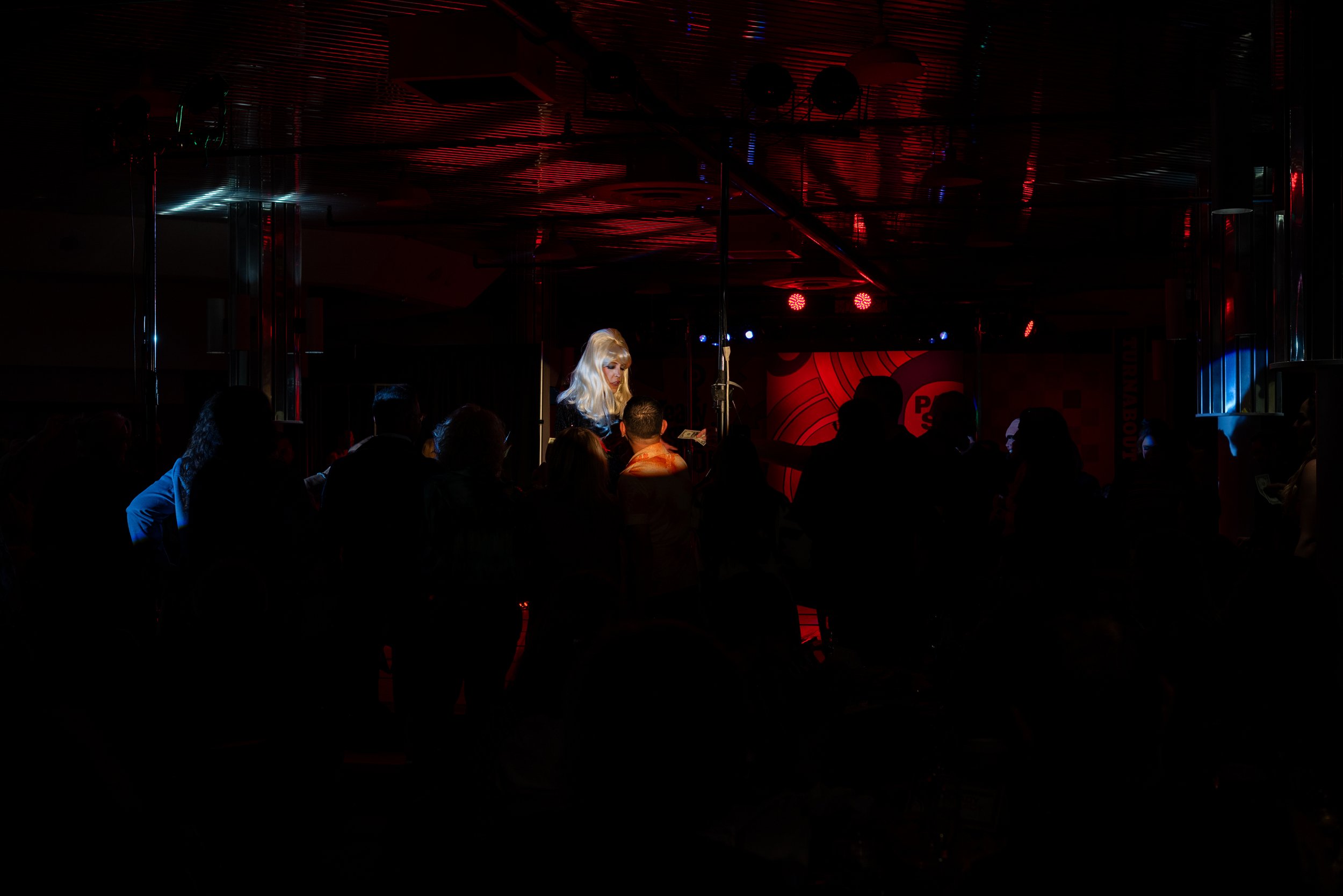 A performer with long blonde hair on stage in an intimate club or concert venue, audience members standing and watching in the dark.