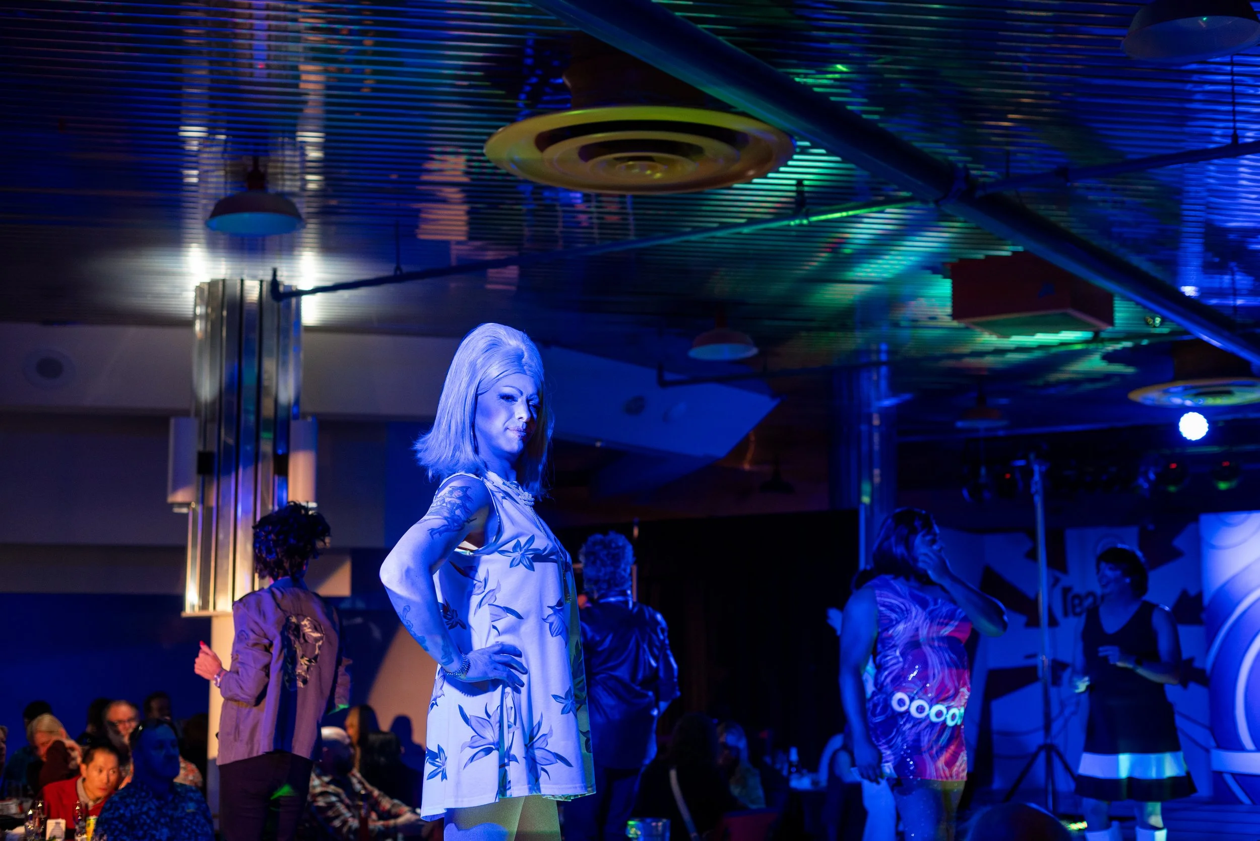 A woman with blonde hair wearing a floral dress stands confidently on stage during a performance, with an audience seated in front of her, under colorful stage lighting.