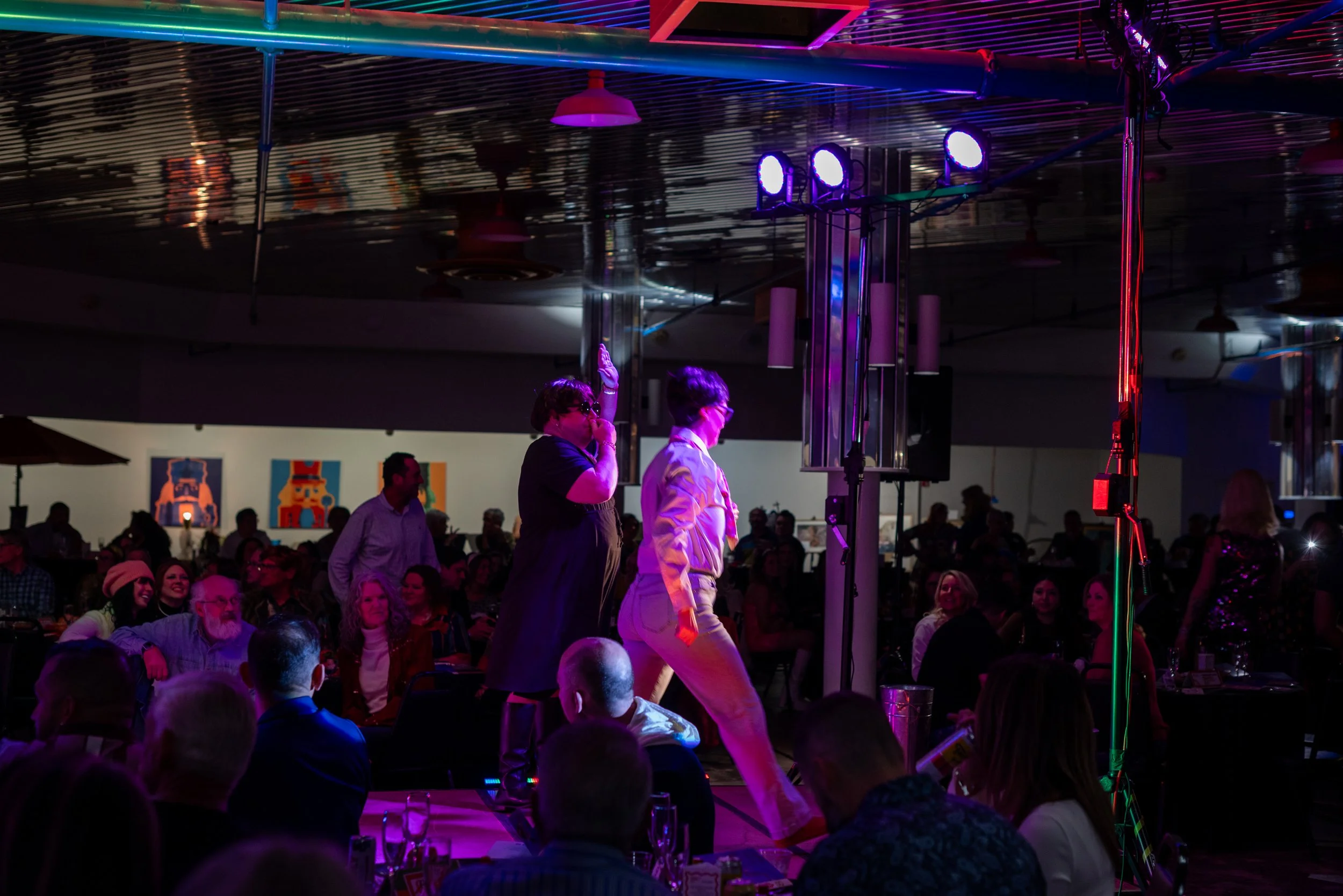 People watching a performance on a stage with purple lighting, some standing and some seated at tables in a dark room with colorful artwork on the walls.