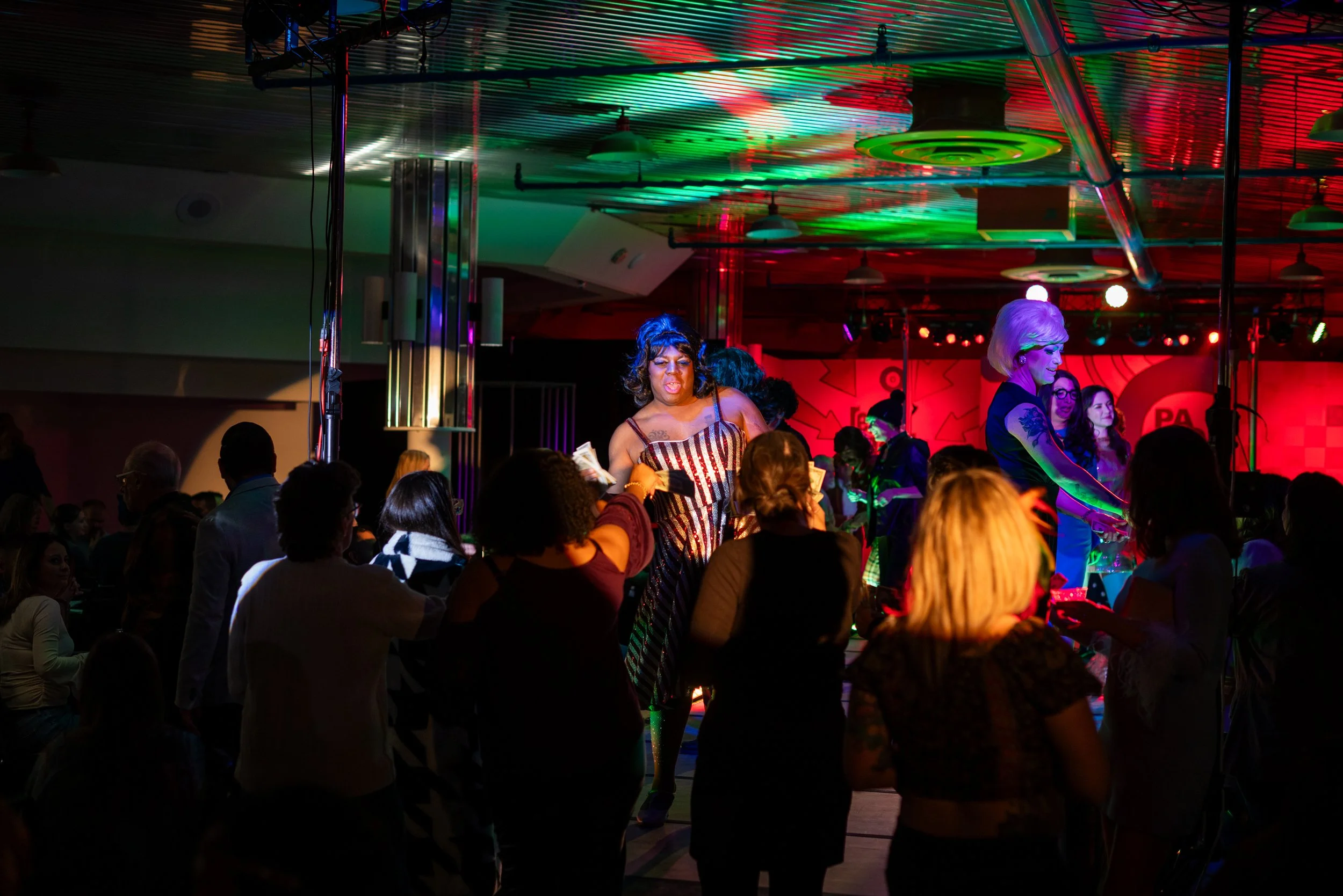 A lively drag show performance at a nightclub with colorful lights, with multiple performers and an audience dancing and enjoying the event.