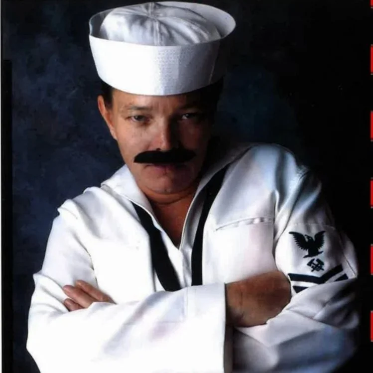 Person dressed as a stereotypical chef with a white chef uniform, hat, and fake mustache, crossing arms and looking serious.