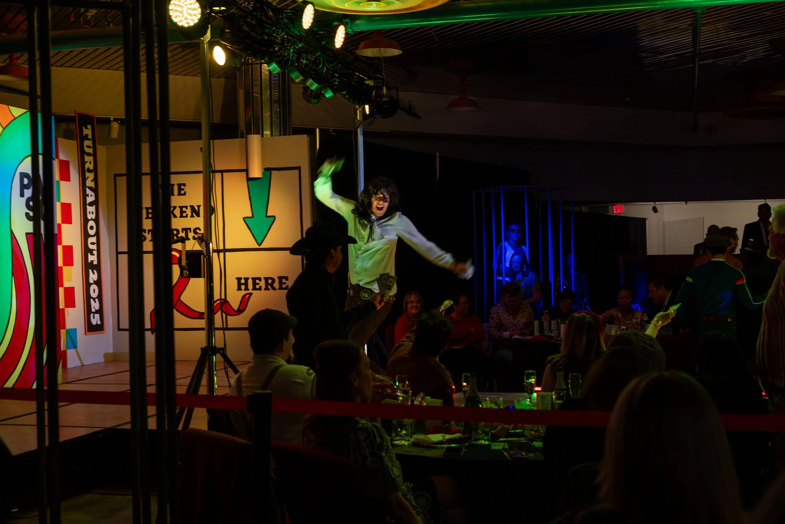 A woman on stage at an event, holding money, with an audience watching. The stage has a colorful backdrop with signs indicating a chicken dance. The setting appears to be lively and festive.