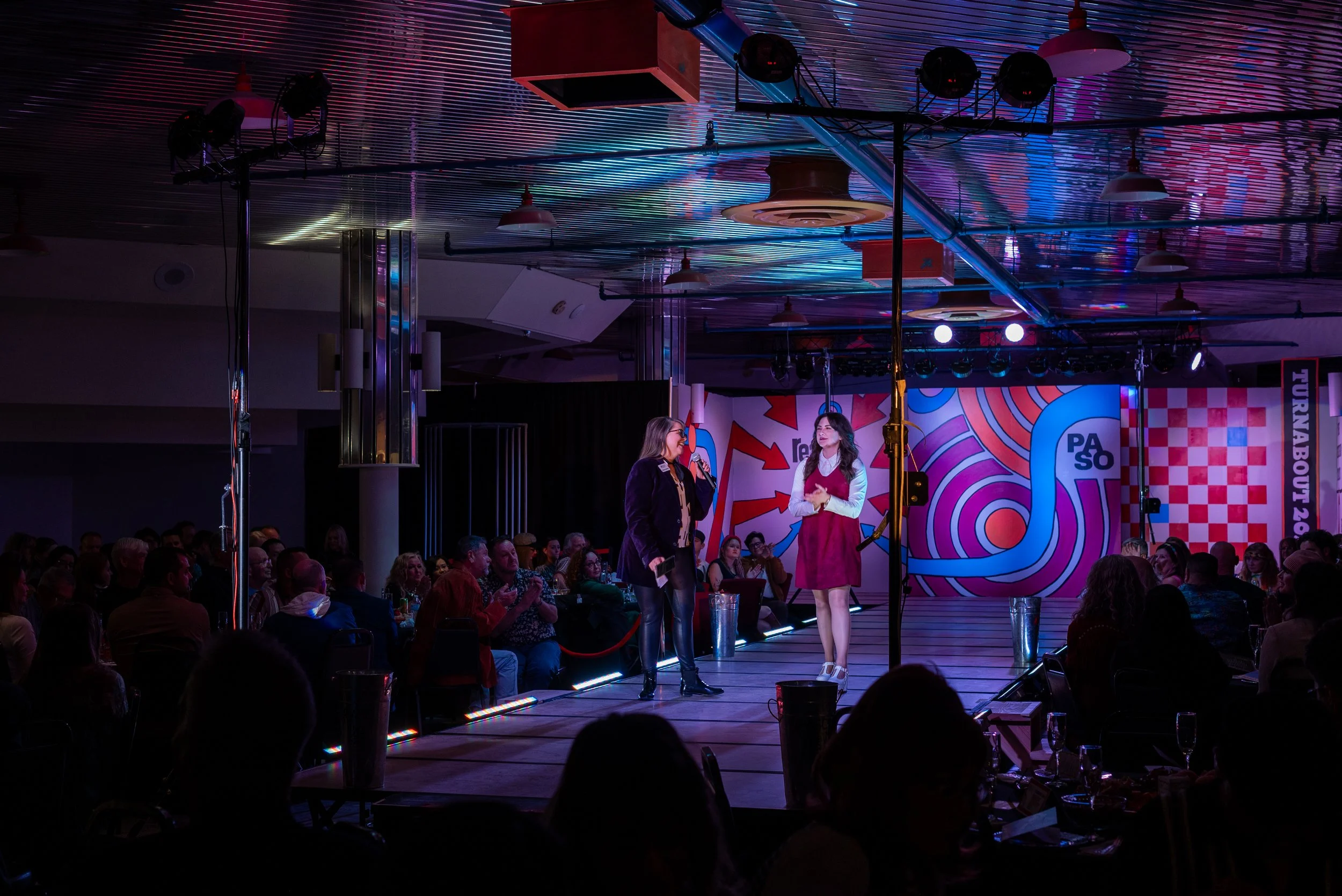 Two women standing on a stage, engaging in a discussion or performance, with a seated audience in a darkened room with colorful lighting and background graphics.