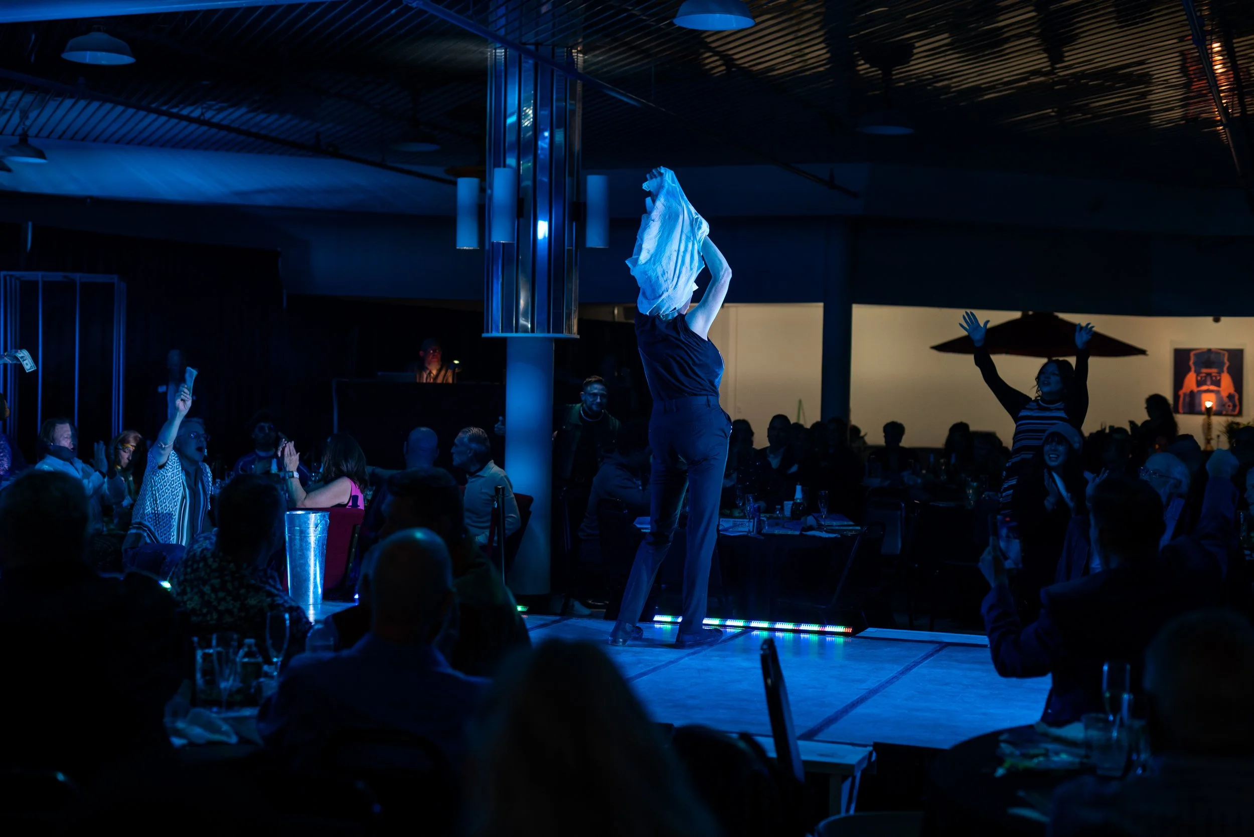 Performer on stage at a dark indoor event, illuminated by blue lights, with audience members seated at tables, some raising hands or taking photos.