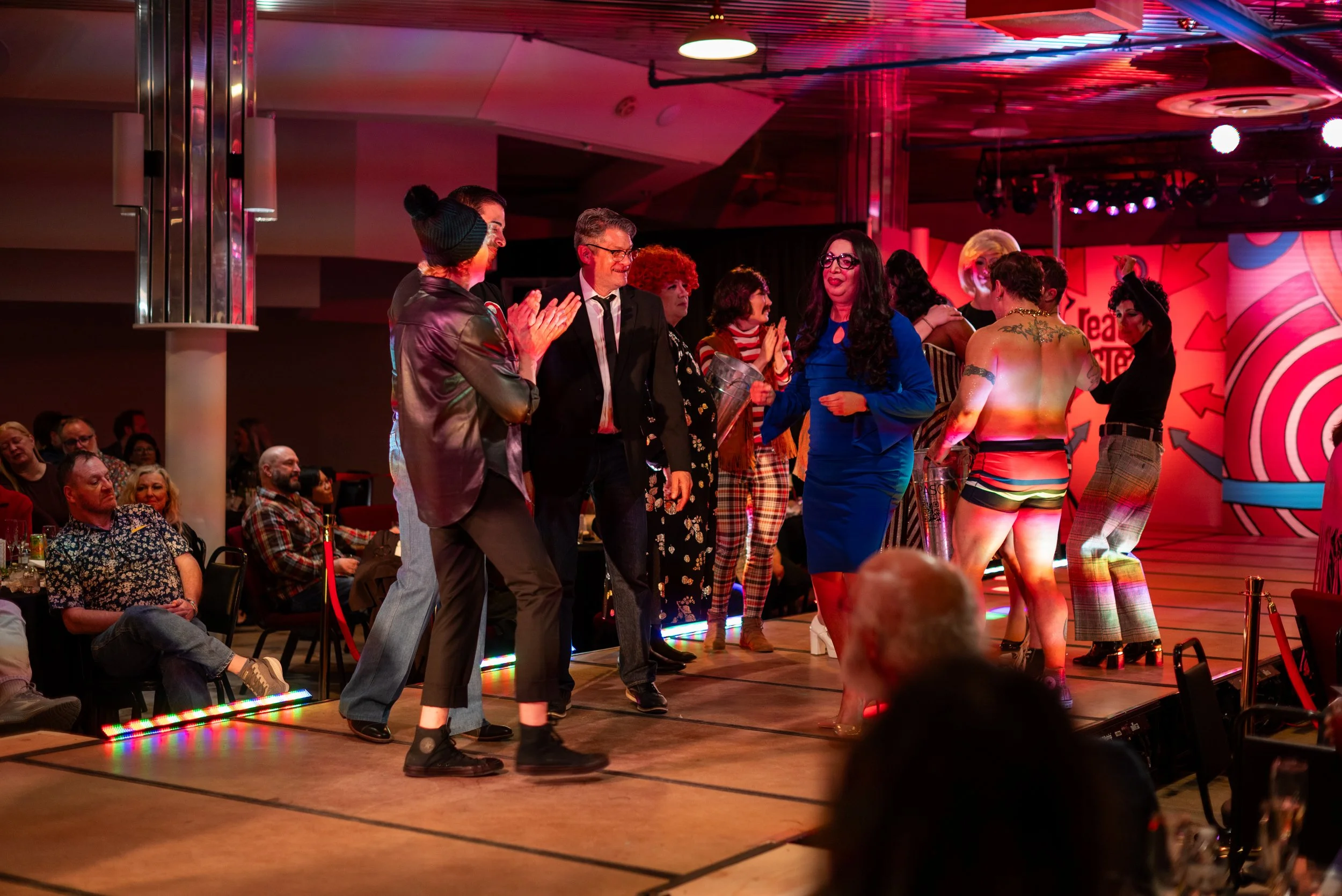 People on stage at a lively event or performance, some clapping and others dancing, with audience watching.