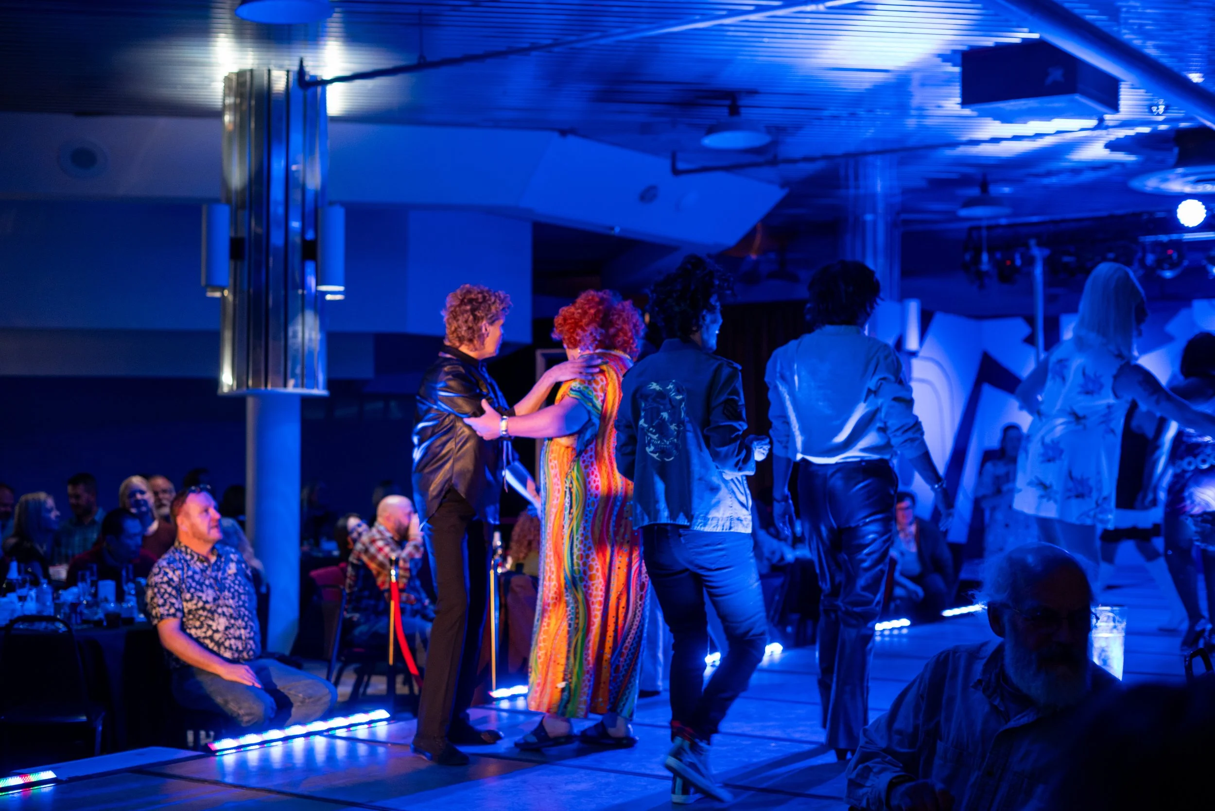 People dancing on stage at a nightclub or event, with audience members seated at tables in the background, under blue lighting.