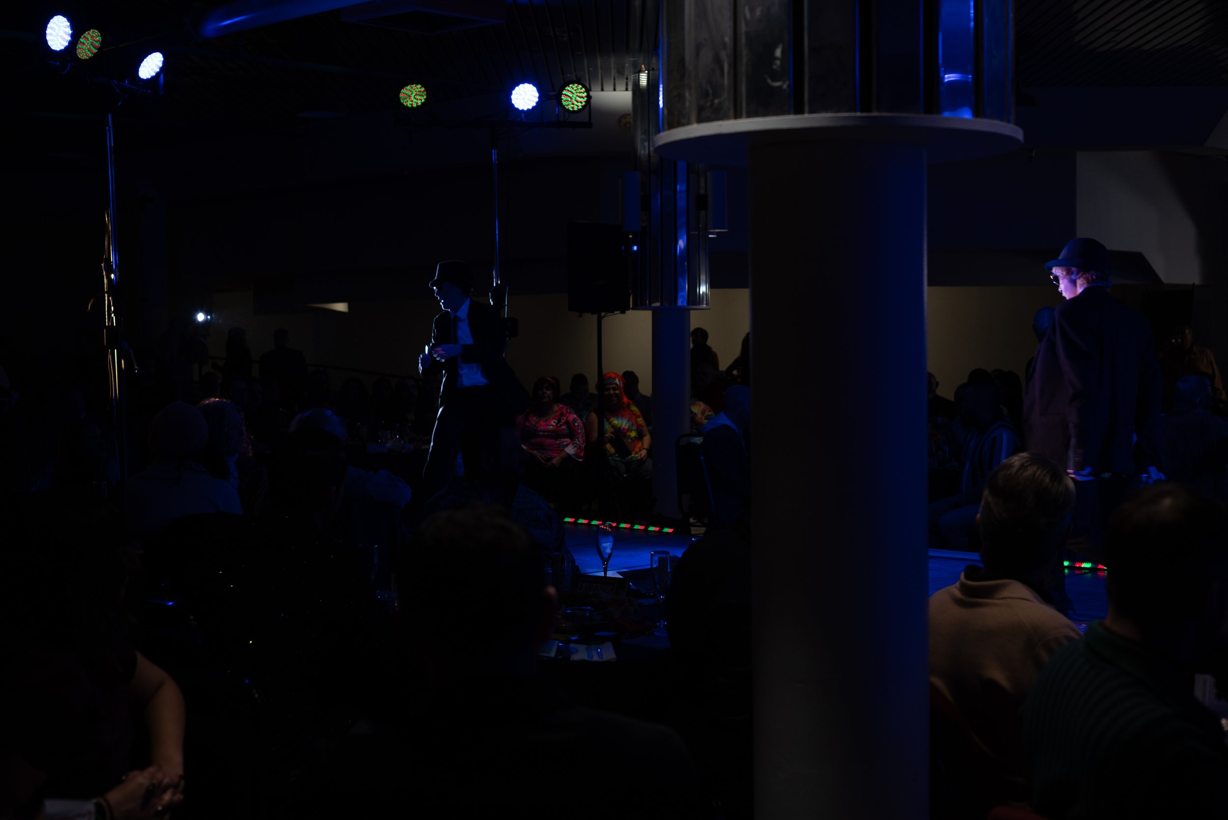 Dimly lit indoor event with a stage, people seated around tables, and two performers on stage illuminated by blue lights.