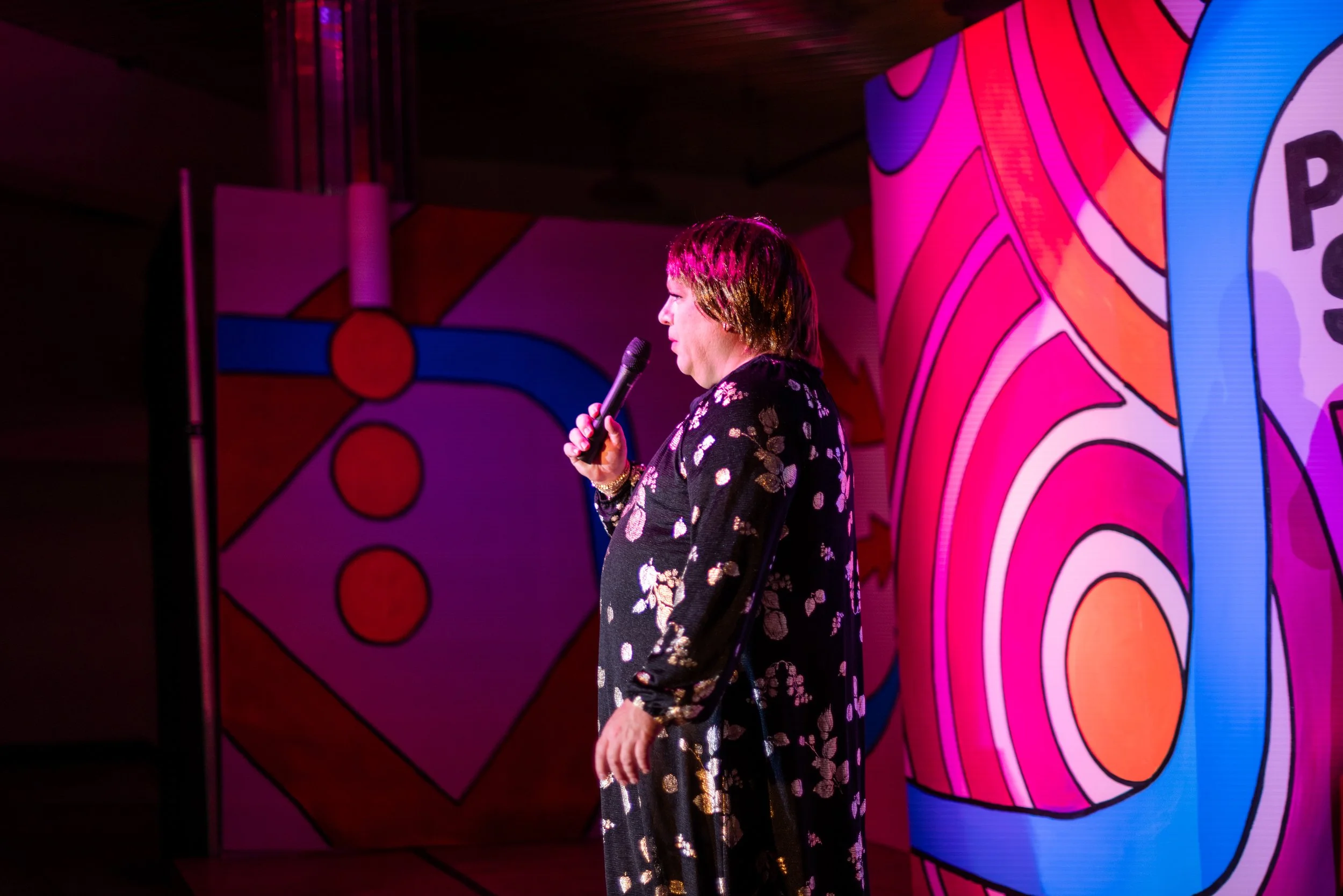 A woman with short hair and dressed in a dark floral jacket is speaking into a microphone on a stage with colorful, abstract art backdrops featuring pink, orange, and blue patterns.