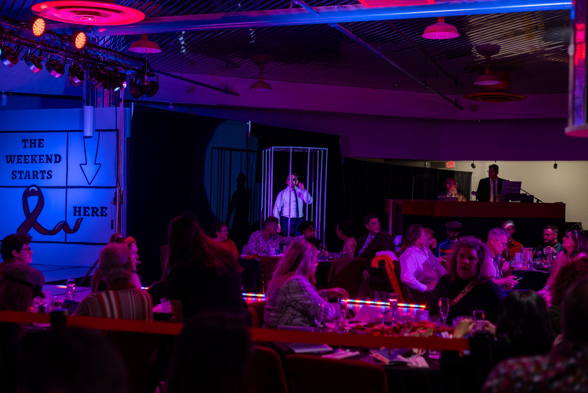 People seated at tables watching a performance in a dimly lit indoor venue with colorful purple and pink lighting. There is a stage with a person inside a cage, and a large sign on the left that says 'The weekend starts here' with an arrow pointing d