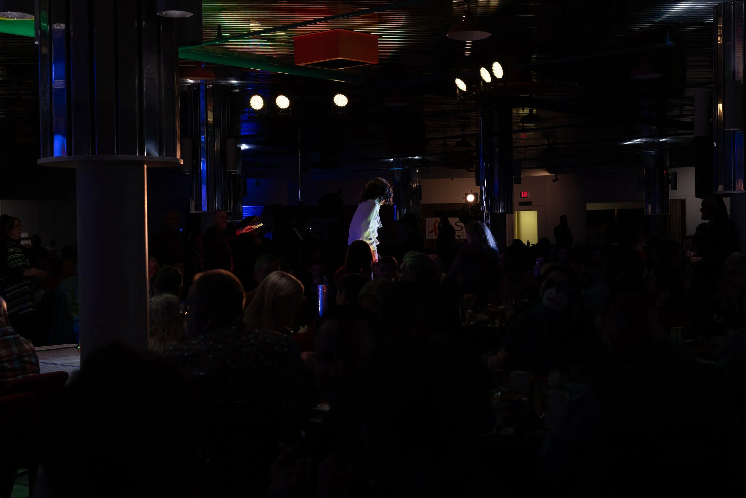 Dimly lit indoor performance venue with a singer on a small stage, surrounded by an audience seated at tables and standing, with colorful ceiling lights.