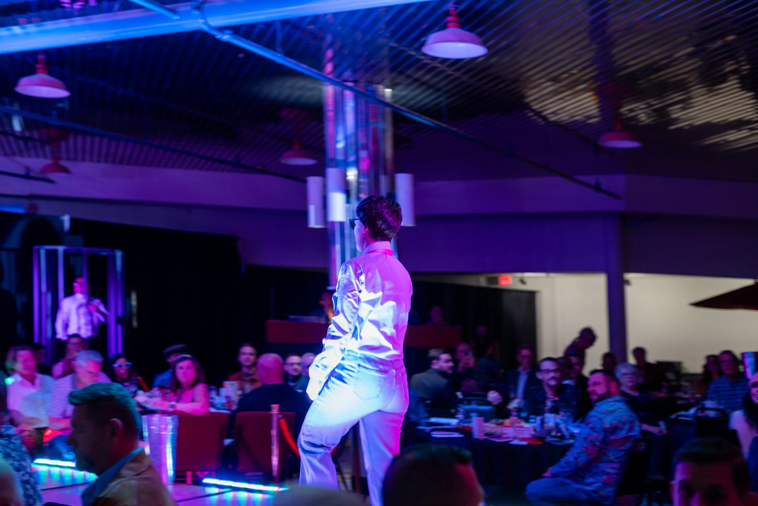 A person in white clothing stands on a stage or platform, facing an audience seated at tables, in a venue with colorful lighting and modern decor.