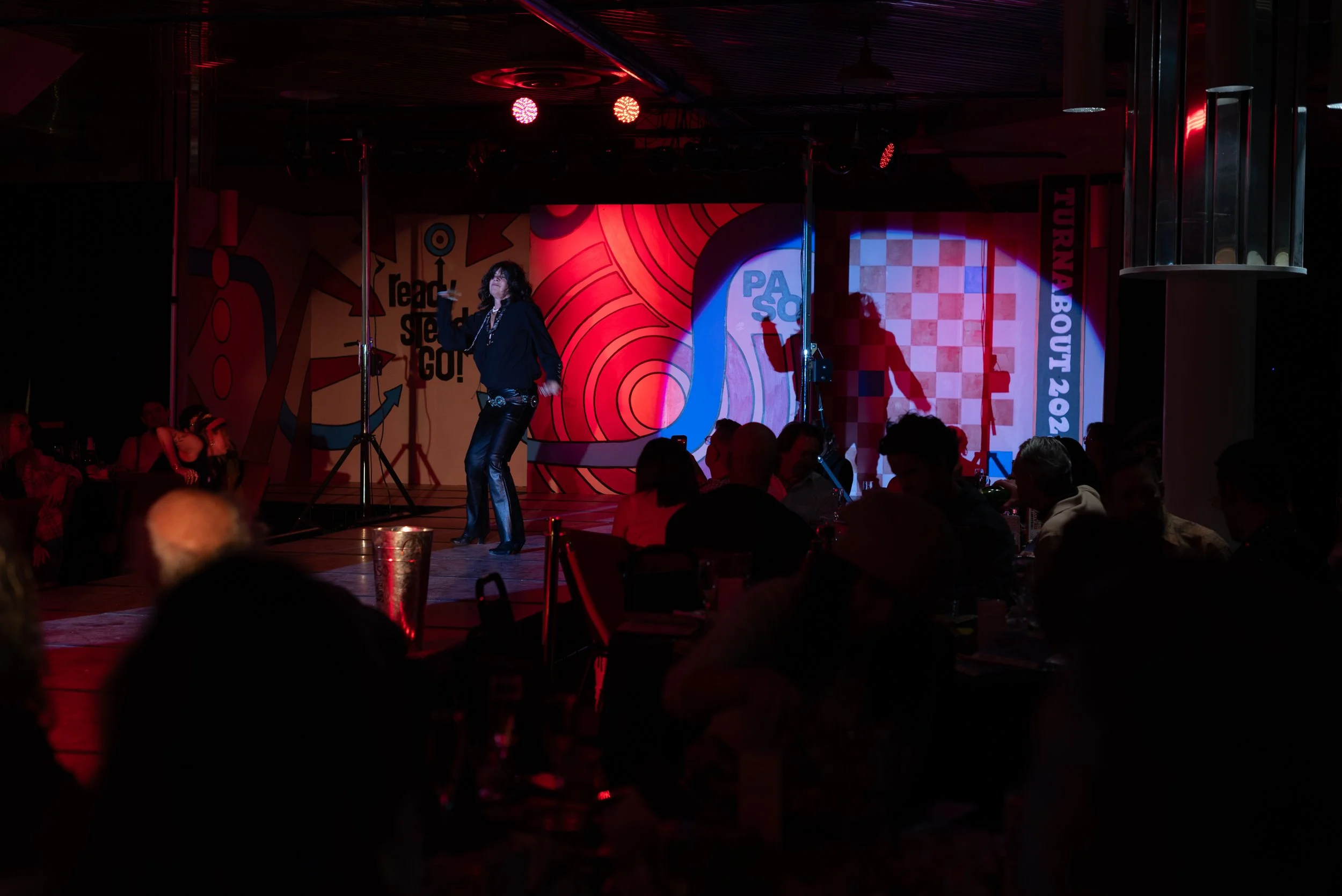 A woman performing on stage at a comedy show or event with the backdrop displaying the text 'PASSPORT' and 'TURN-BOUT 2023'. The stage has colorful, artistic graphics and audience members seated in front of her in a darkened room.