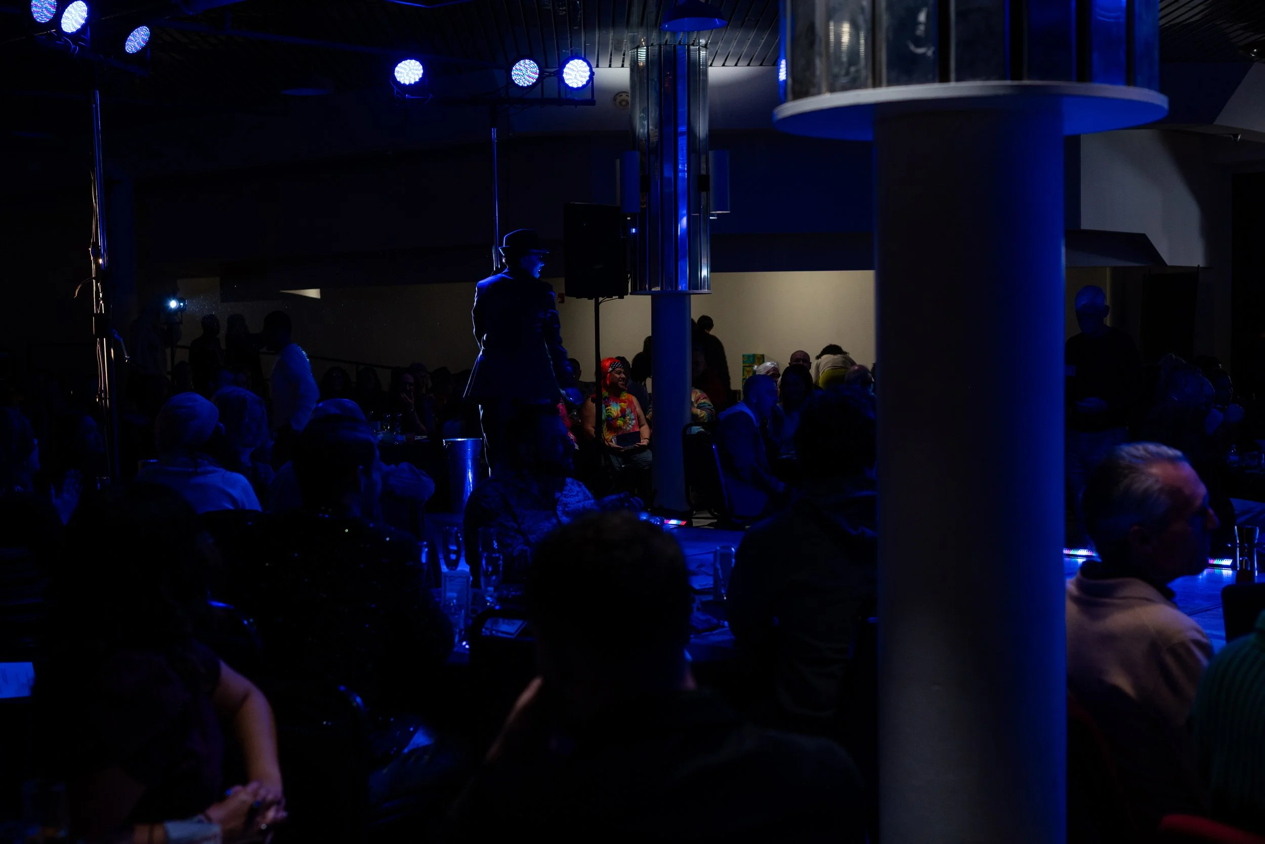 A dark indoor event with people seated at tables, illuminated by blue lighting, with a person in a hat and suit standing and speaking into a microphone in the center.