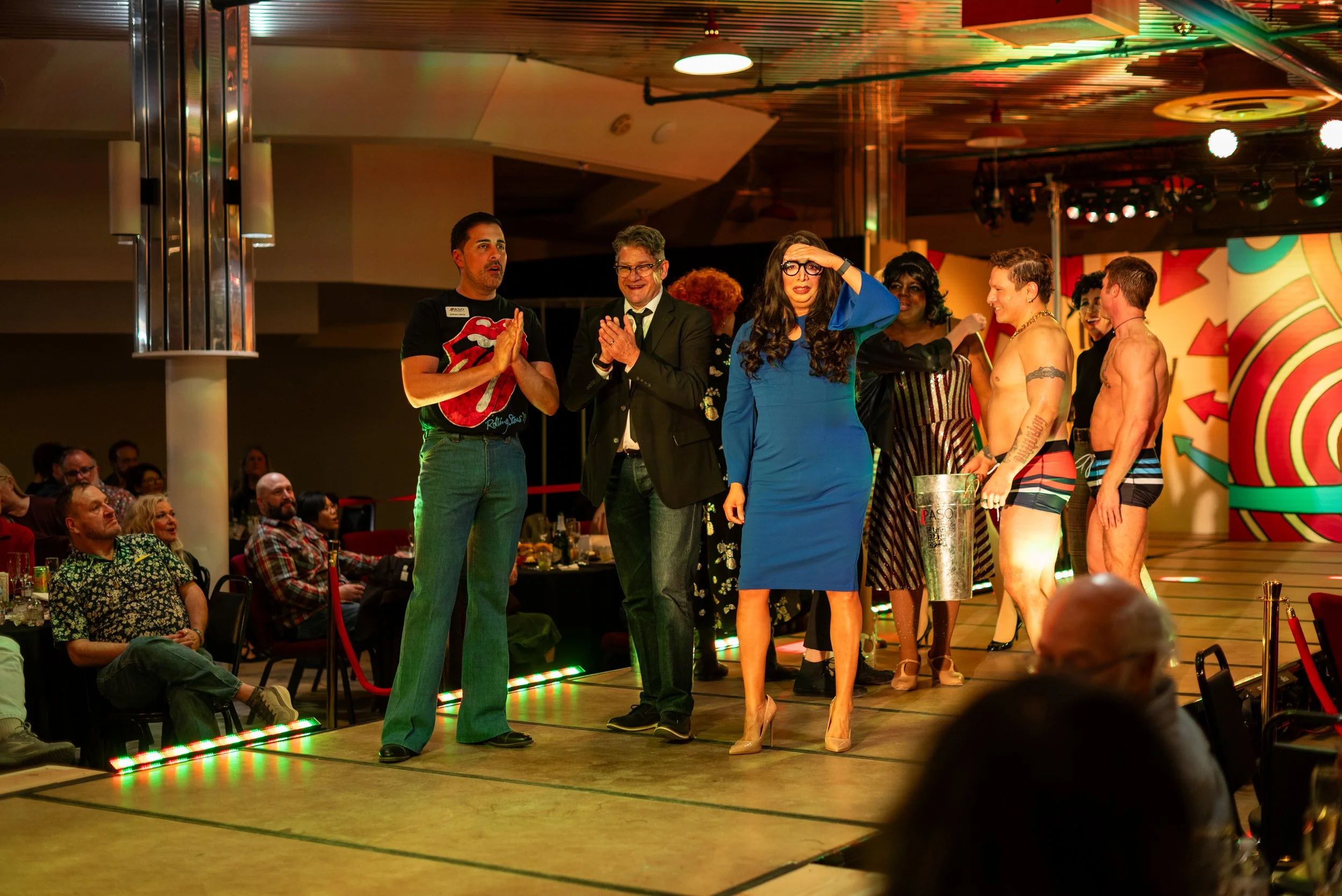 Group of people on a stage at a lively event, some in colorful costumes, others in formal attire, with an audience watching in the background.