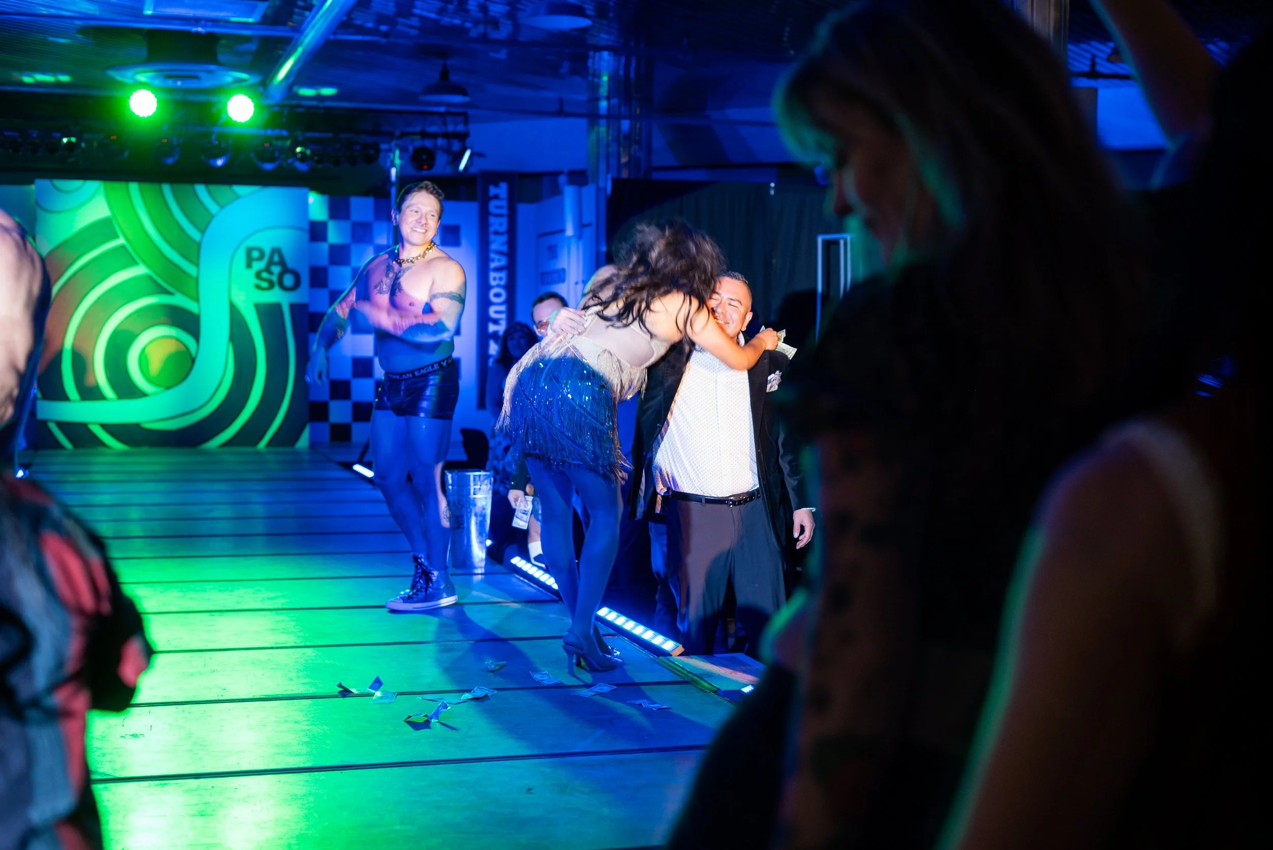People dancing and socializing at a nightclub or party with a brightly lit stage and colorful background.