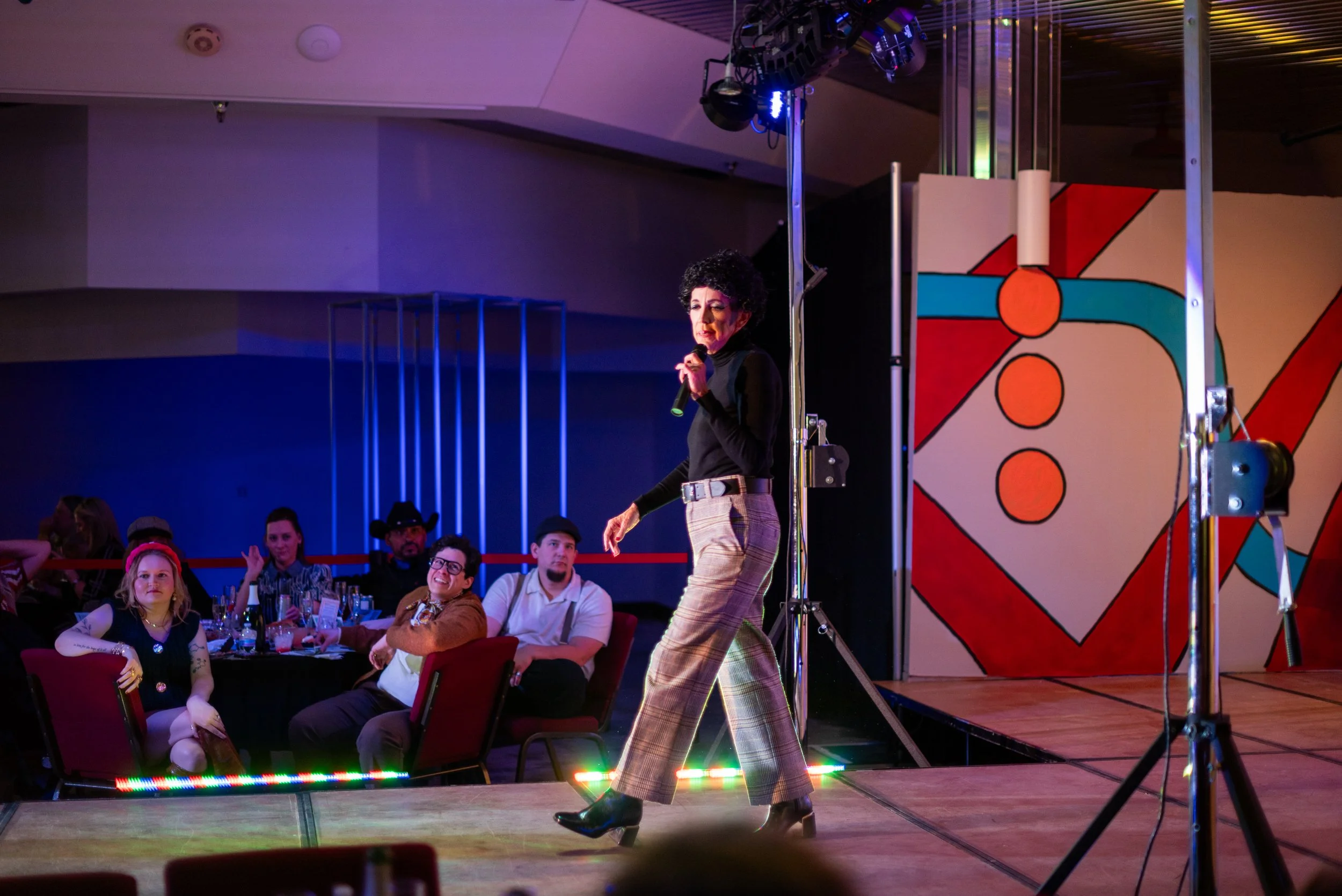 A woman with curly black hair dressed in black top and plaid pants walking on a stage holding a microphone at a social event, with seated guests watching her.