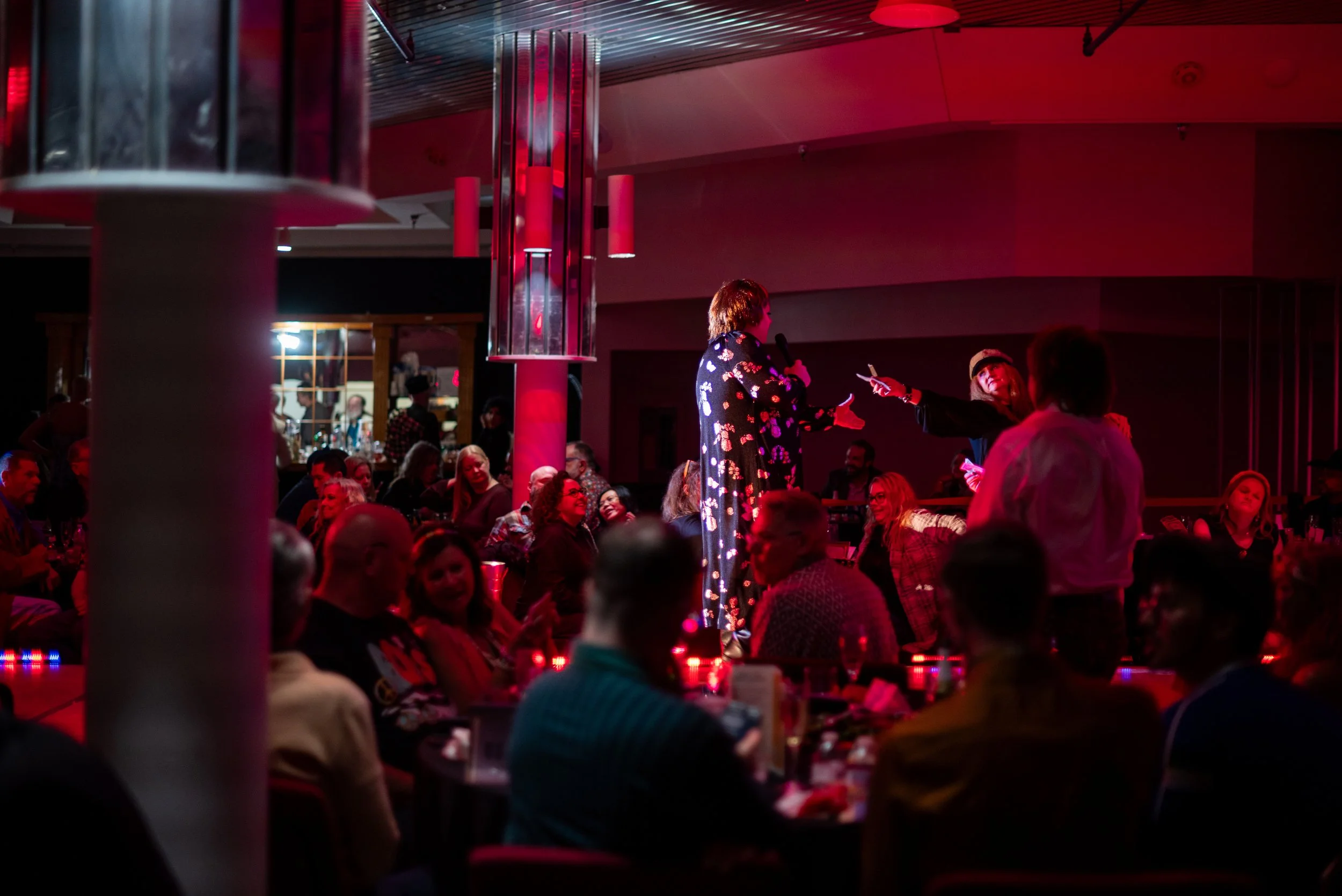 A lively indoor event with a woman on stage speaking into a microphone, surrounded by seated attendees, illuminated with red and purple lighting.