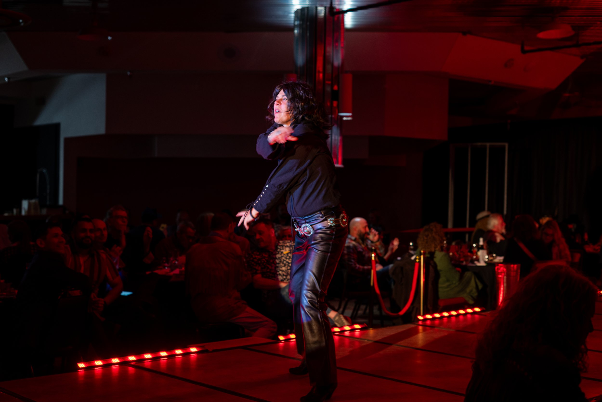 Performer with curly hair singing or speaking on stage in front of audience at a nightclub or concert venue, with red lighting and seated spectators