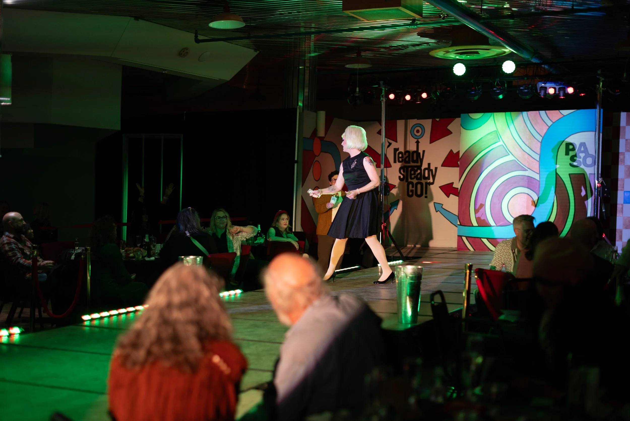 Woman on stage at a comedy or entertainment show, with a colorful backdrop that says 'Ready Steady GO!', audience seated at tables in a dimly lit atmosphere.