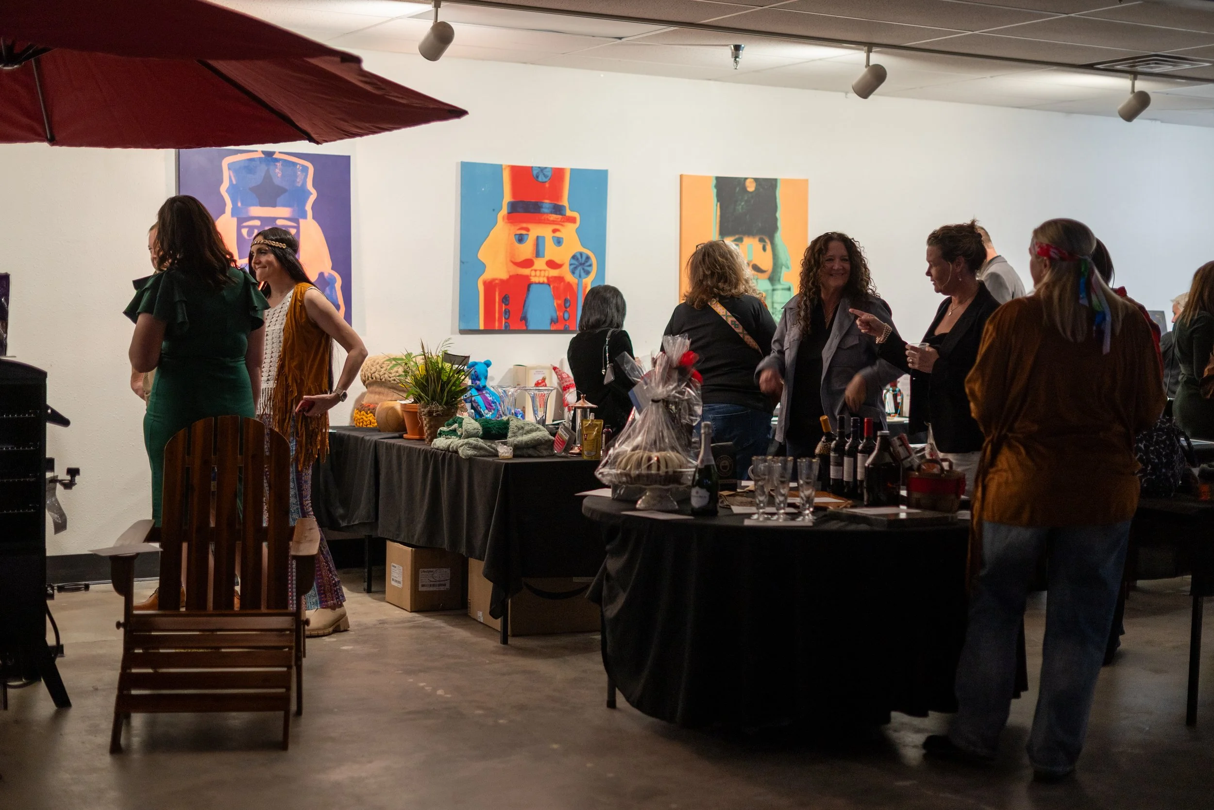 People mingling at an art gallery opening, with colorful artwork on the white walls, tables displaying food and drinks, and a red umbrella in the foreground.