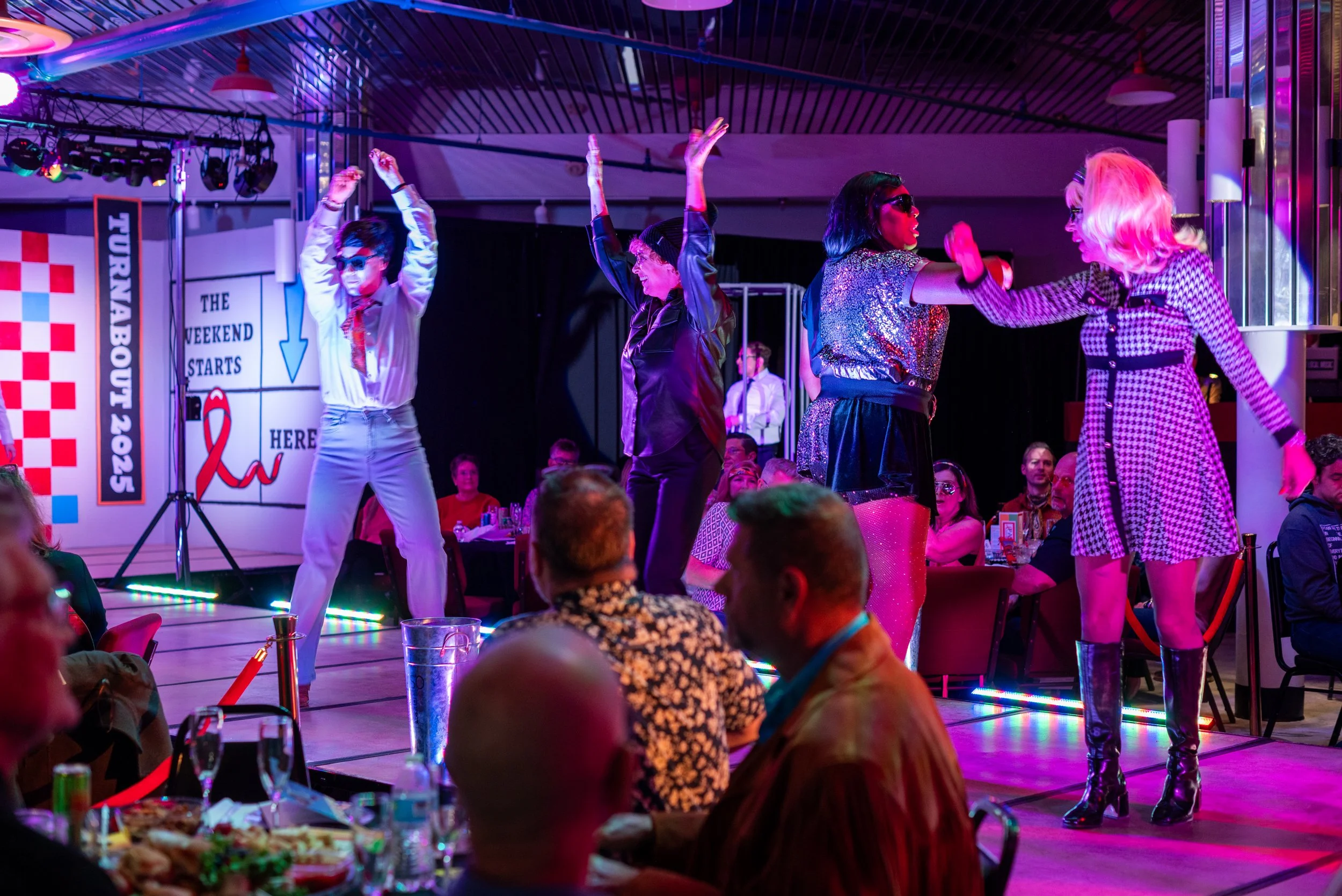 People performing a dance on stage at a party or event, with audience seated at tables watching.