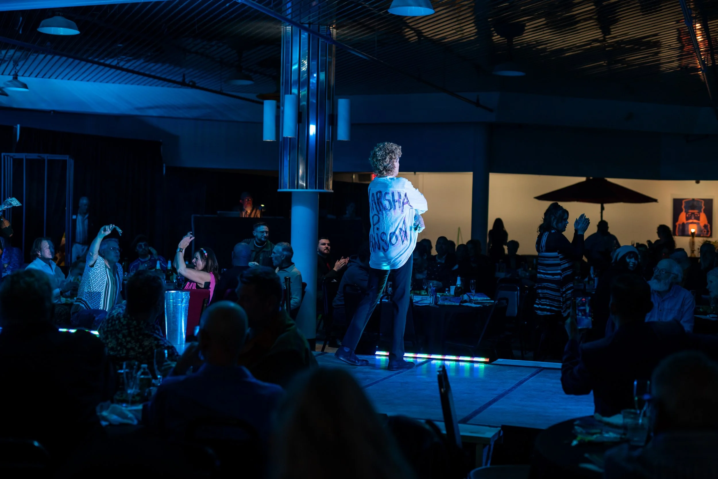 A performer on stage at a dark indoor event, wearing a white shirt with writing on the back, facing an audience seated at tables, some taking photos or clapping, under colorful lighting.