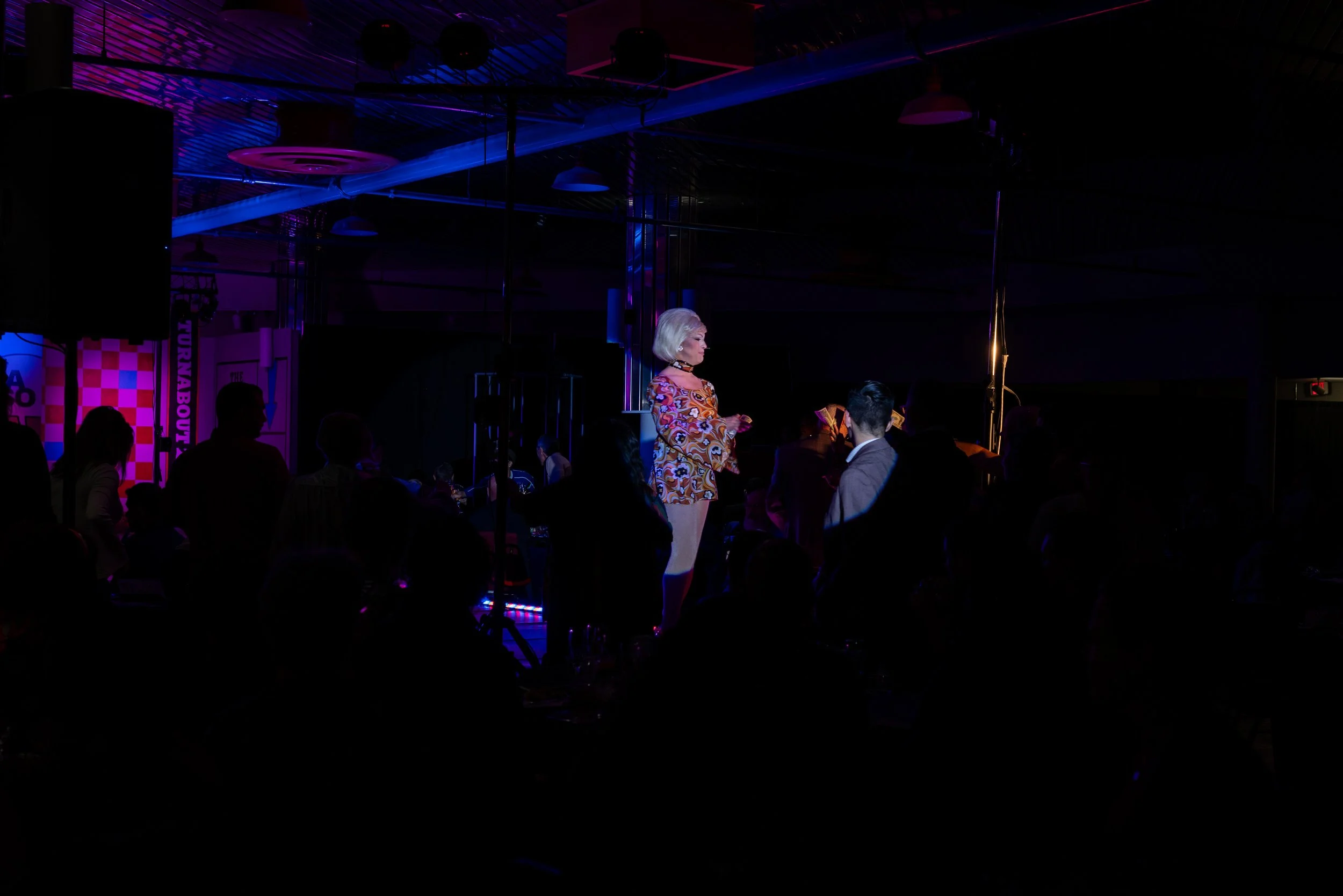 A performer with platinum blonde hair singing on a stage illuminated by colorful lights, while an audience watches in a dark venue.