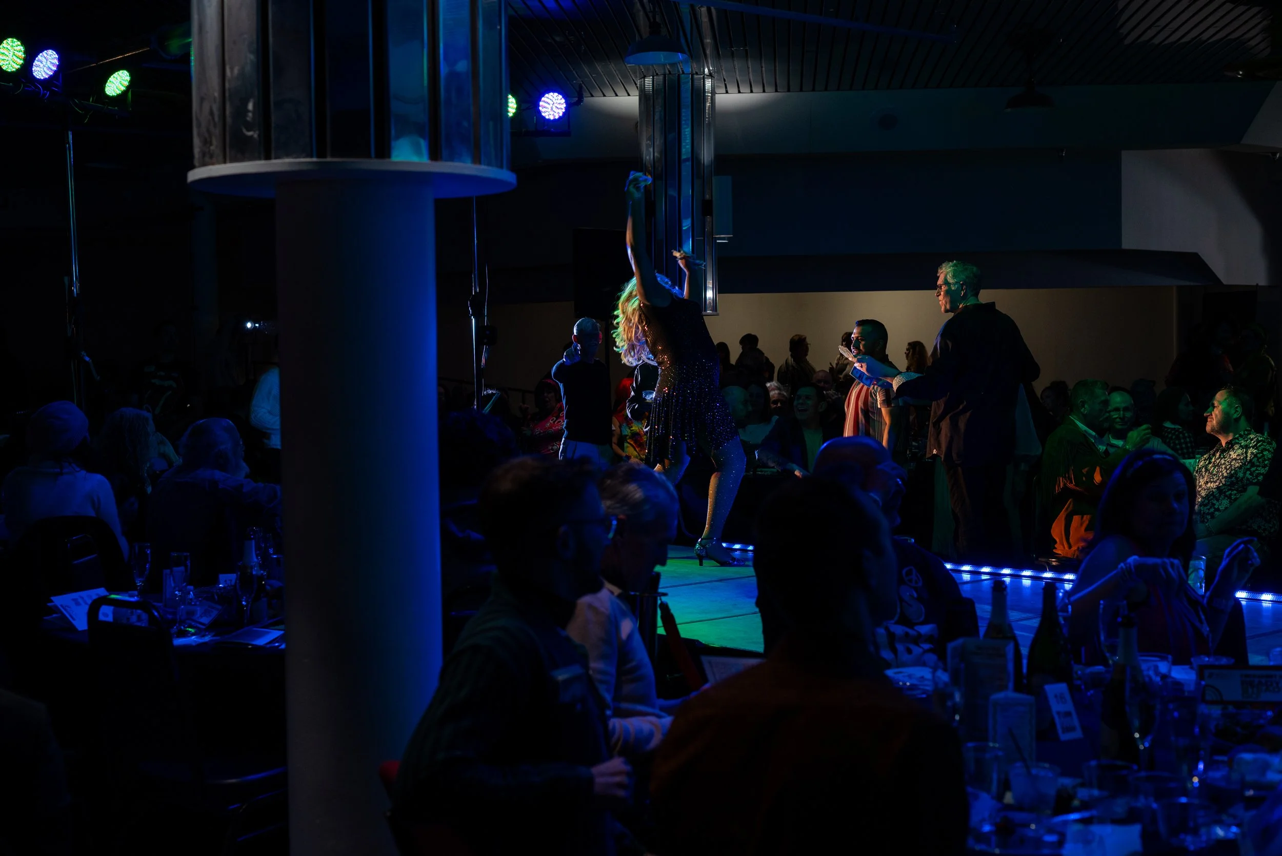 A woman dancing on a stage with her arms raised, surrounded by people seated at tables, in a dark, colorful nightclub or event setting with stage lights.