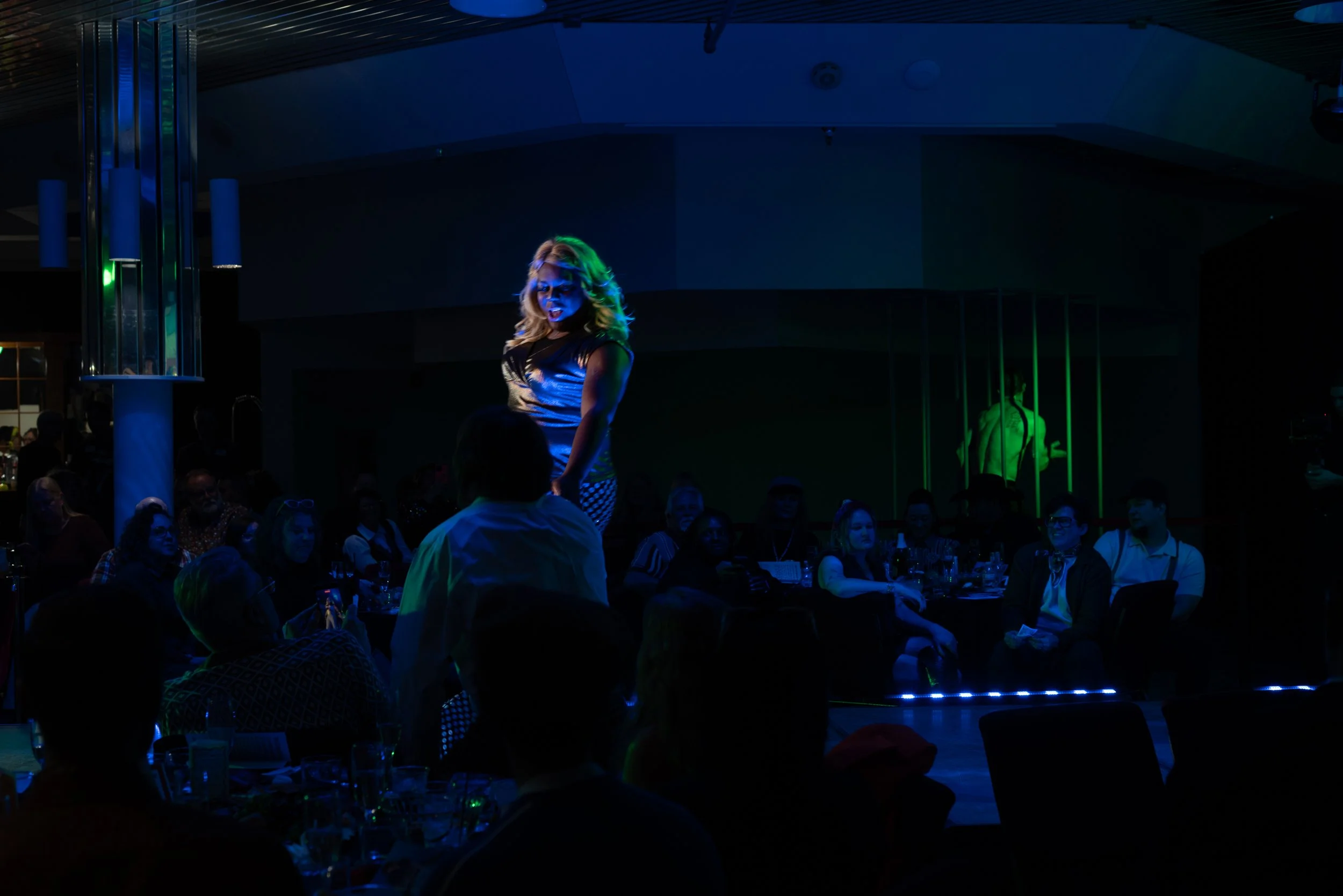 A woman performing on a stage in a dark, nightclub setting with an audience watching. The woman is illuminated by colorful stage lights, and a man is speaking or singing into a microphone in front of her.