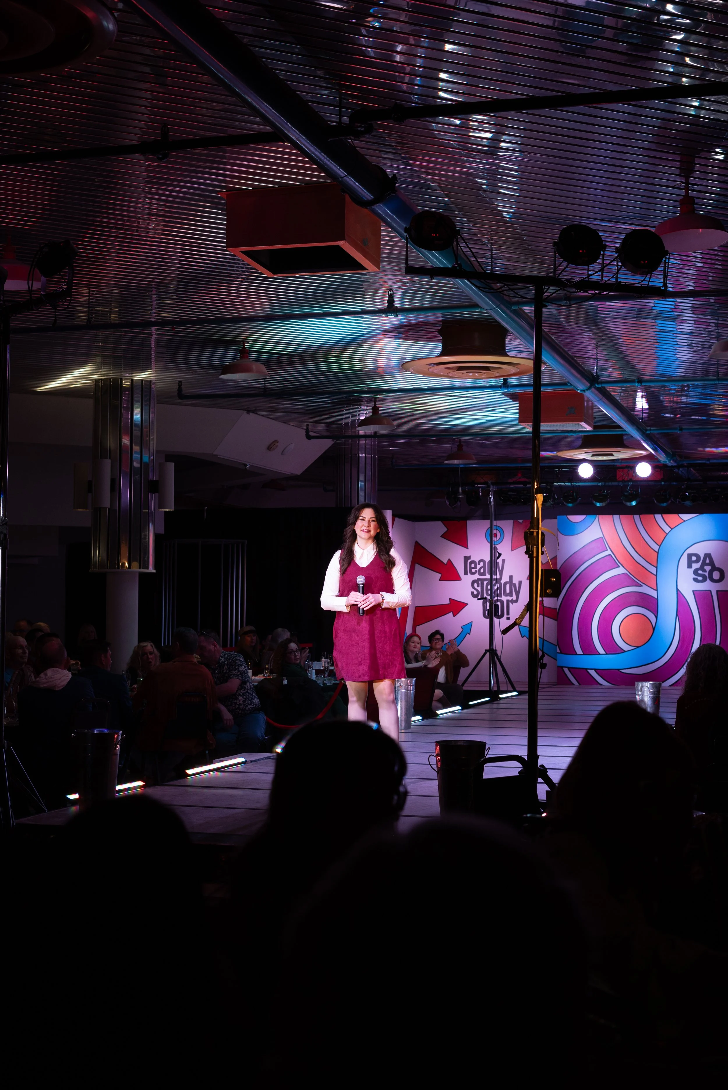 A woman standing on a stage holding a microphone during a comedy show or presentation, with an audience seated in front of her and colorful, vibrant backgrounds on the walls.