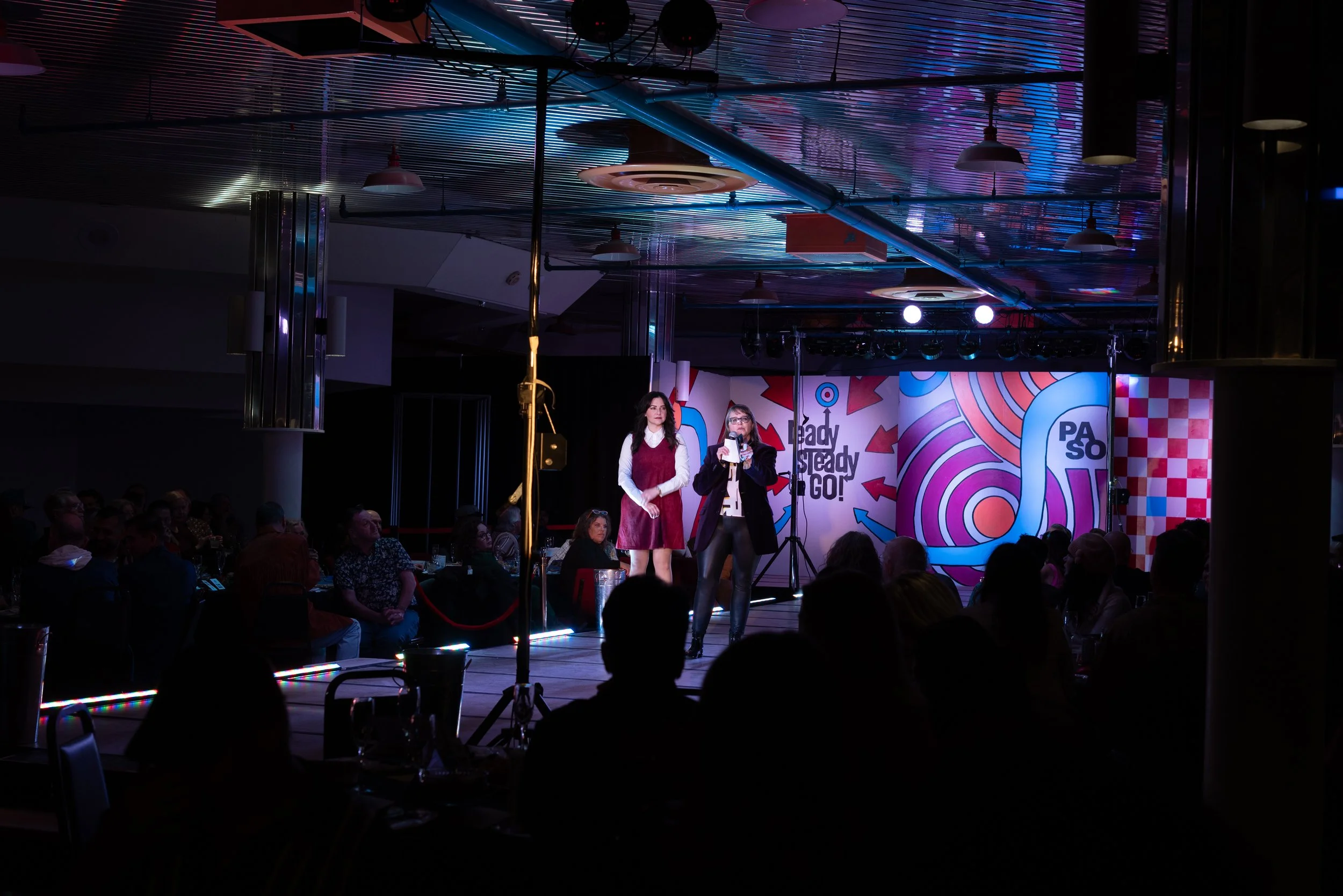 Two women standing on stage in front of a seated audience, with colorful abstract backdrop and stage lighting, during an event or presentation.