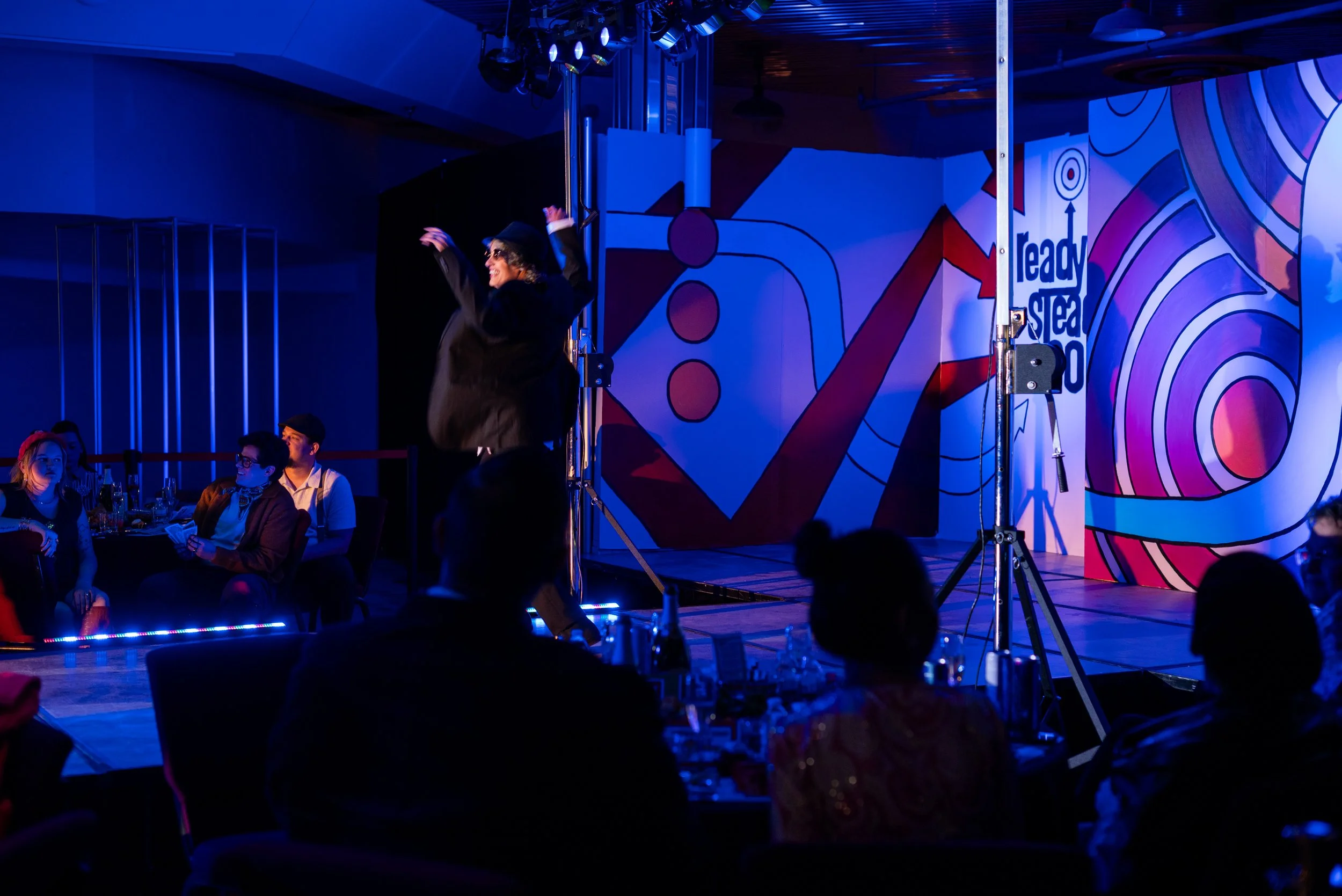 A performer on stage dancing at an event with guests seated around. The stage has colorful abstract art and the words "ready to steal the show" on the wall.