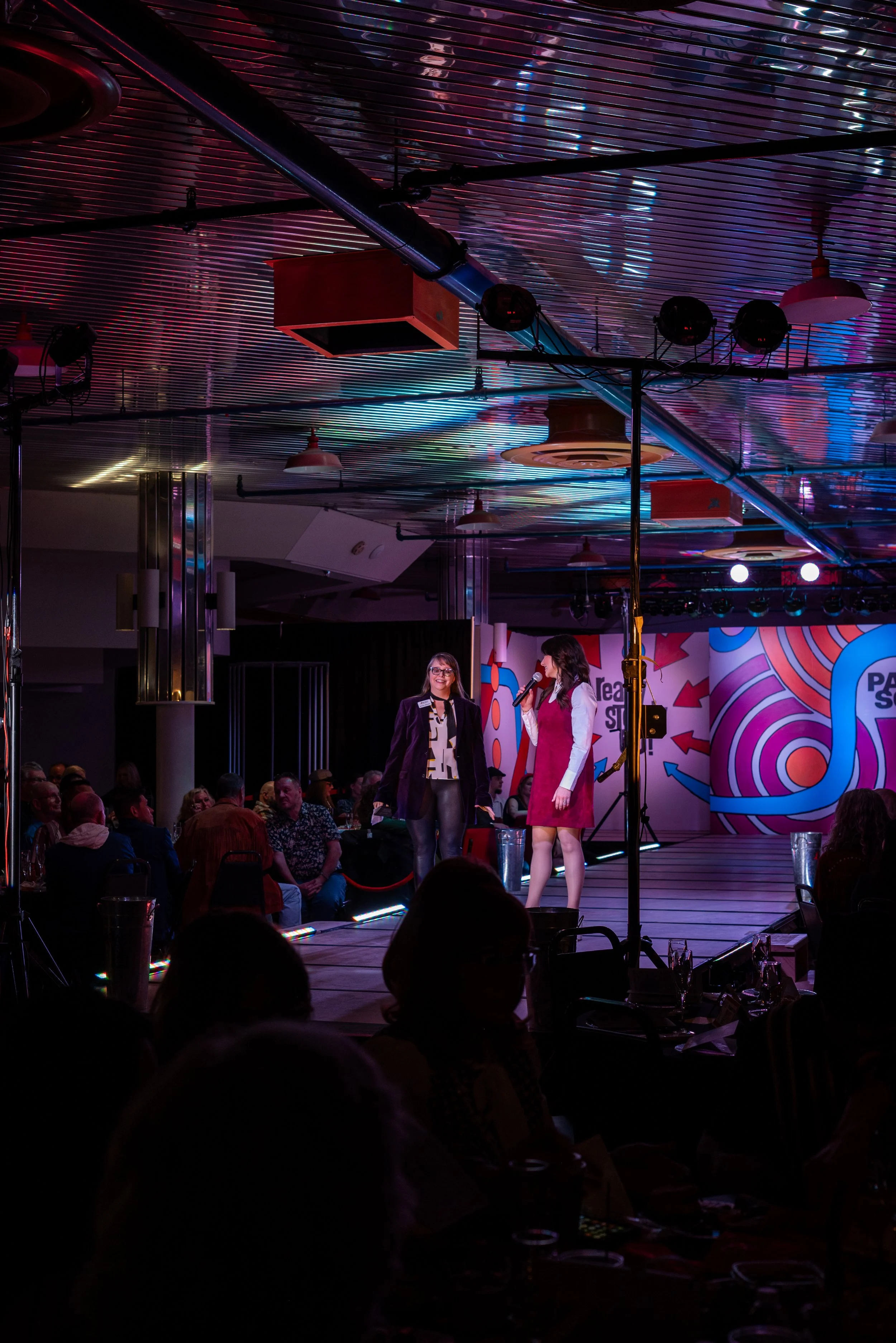 Two women on stage at an event, with an audience seated in front, colorful abstract background, and stage lighting.