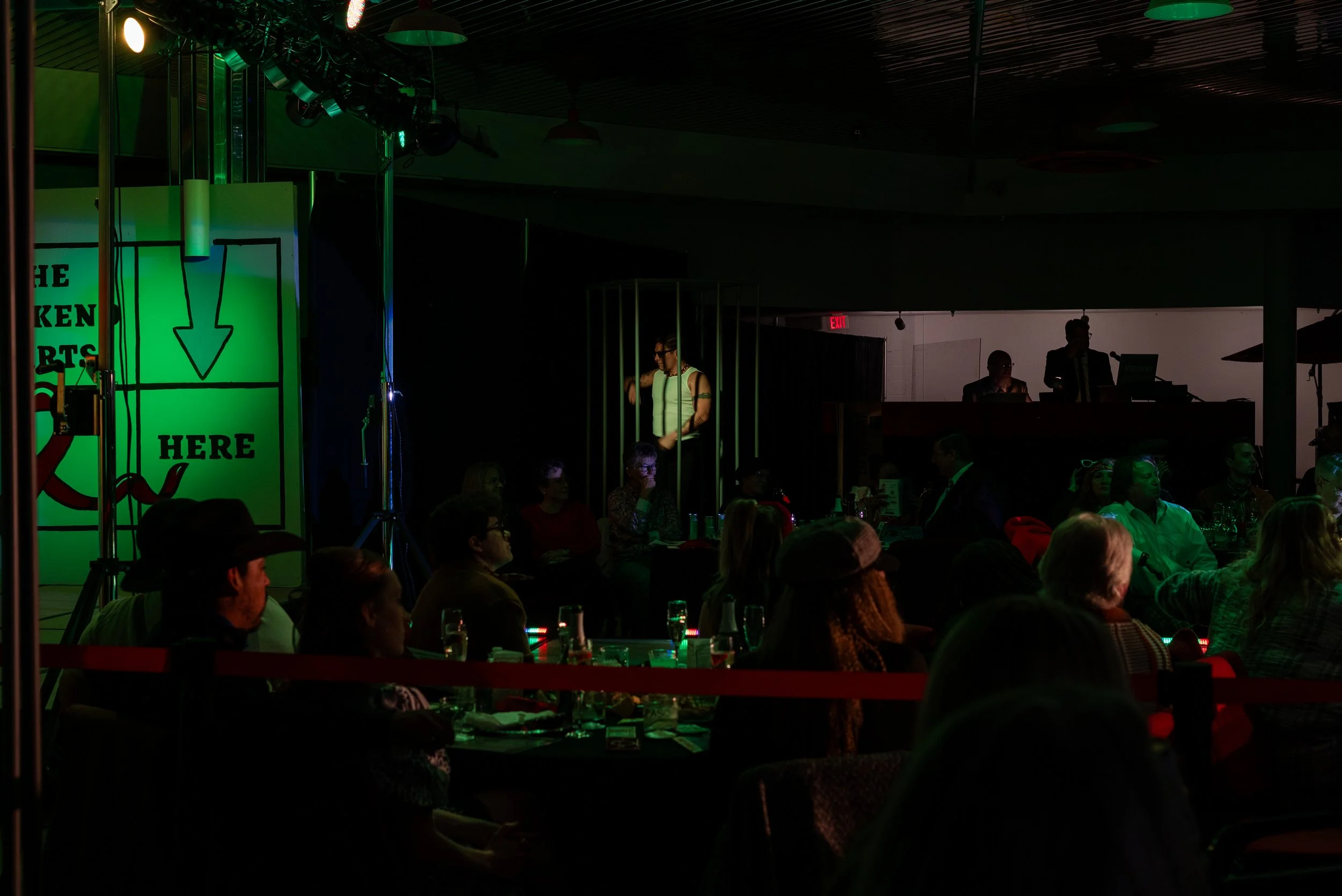 Dark indoor comedy show with audience seated and illuminated stage with a large sign reading 'Here' with an arrow pointing down.