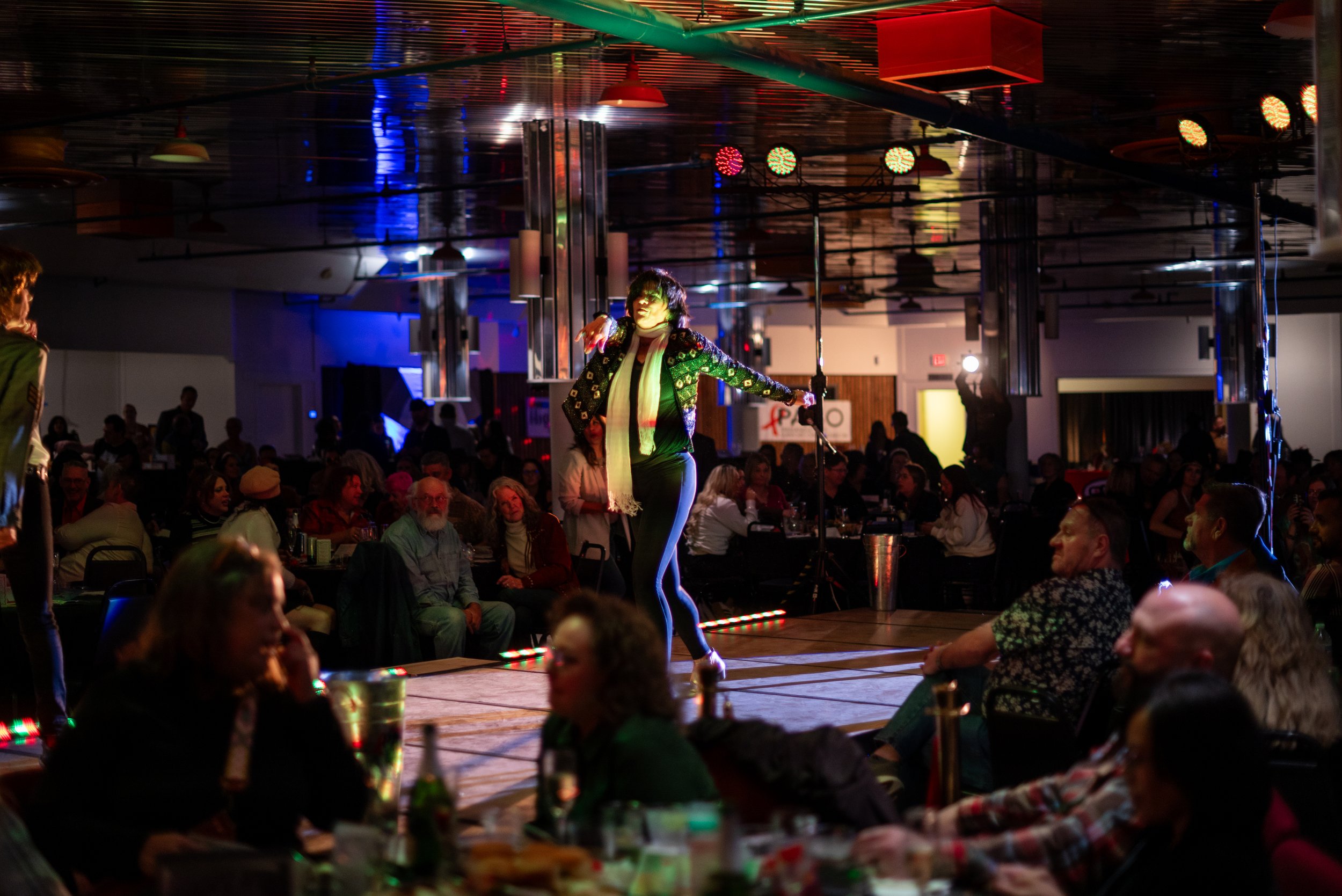 A performer singing on a stage with a microphone in hand, surrounded by an audience in a dimly lit venue with colorful stage lights.