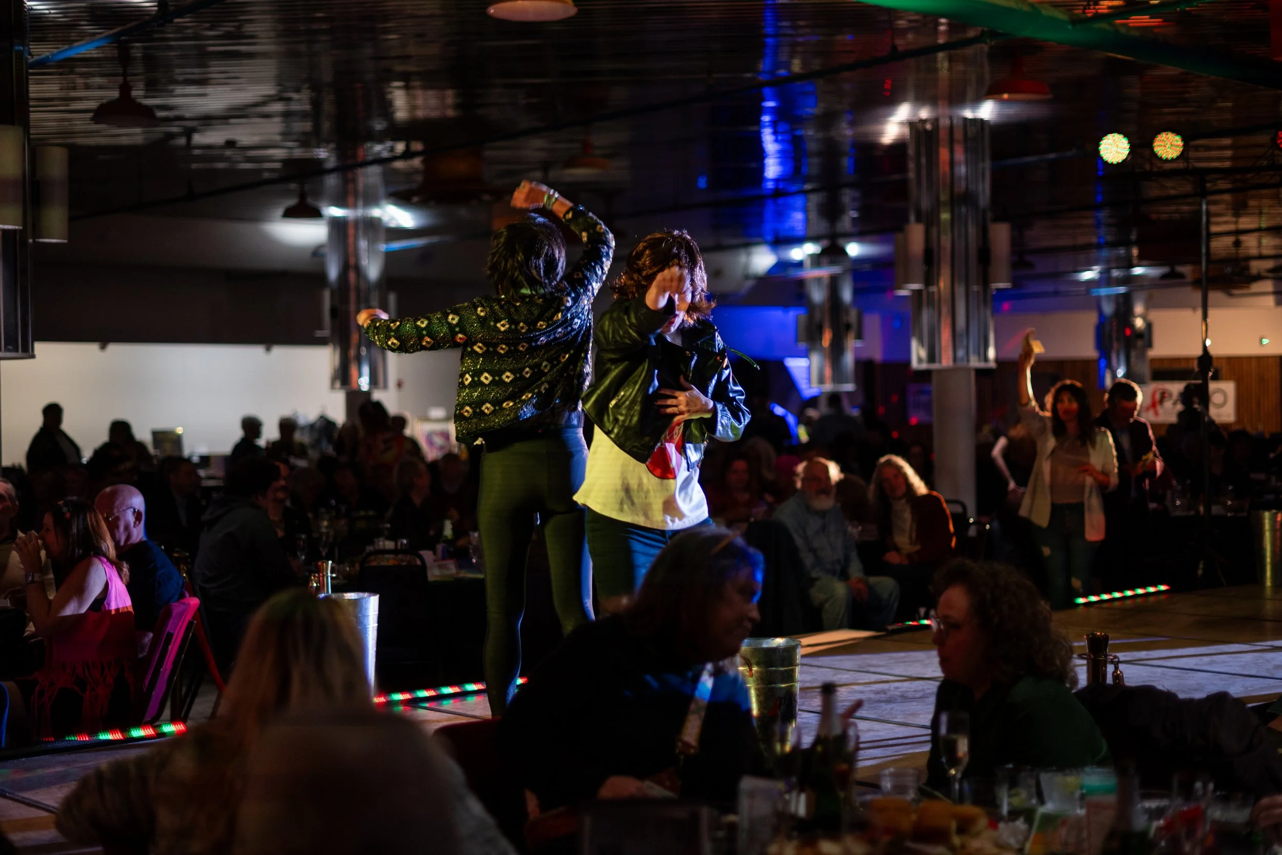 A dance floor in a dimly lit nightclub with two women dancing in the foreground, surrounded by seated and standing people, with colorful lights and a lively atmosphere.