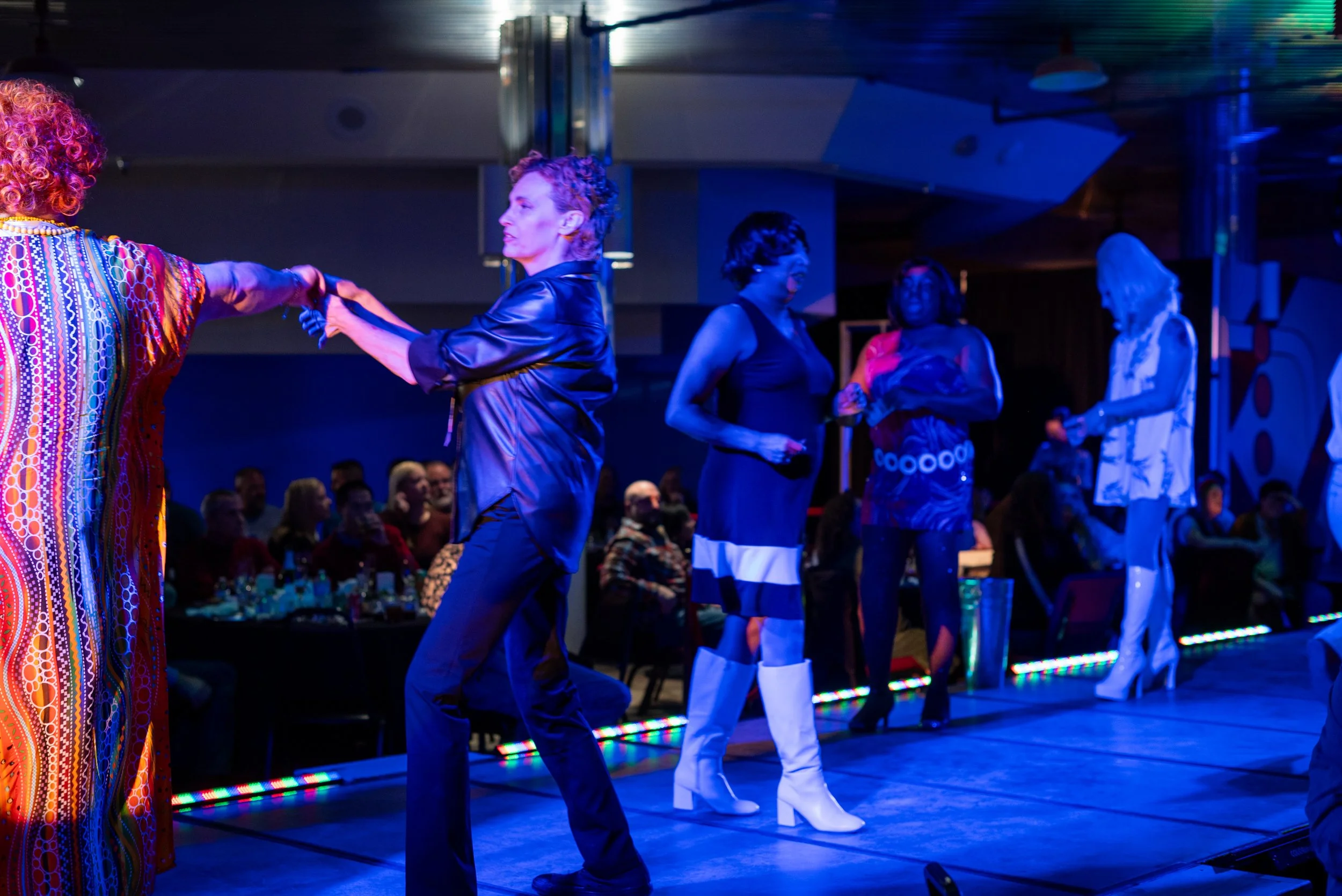 Group of women line dancing on stage at night, with audience in background.