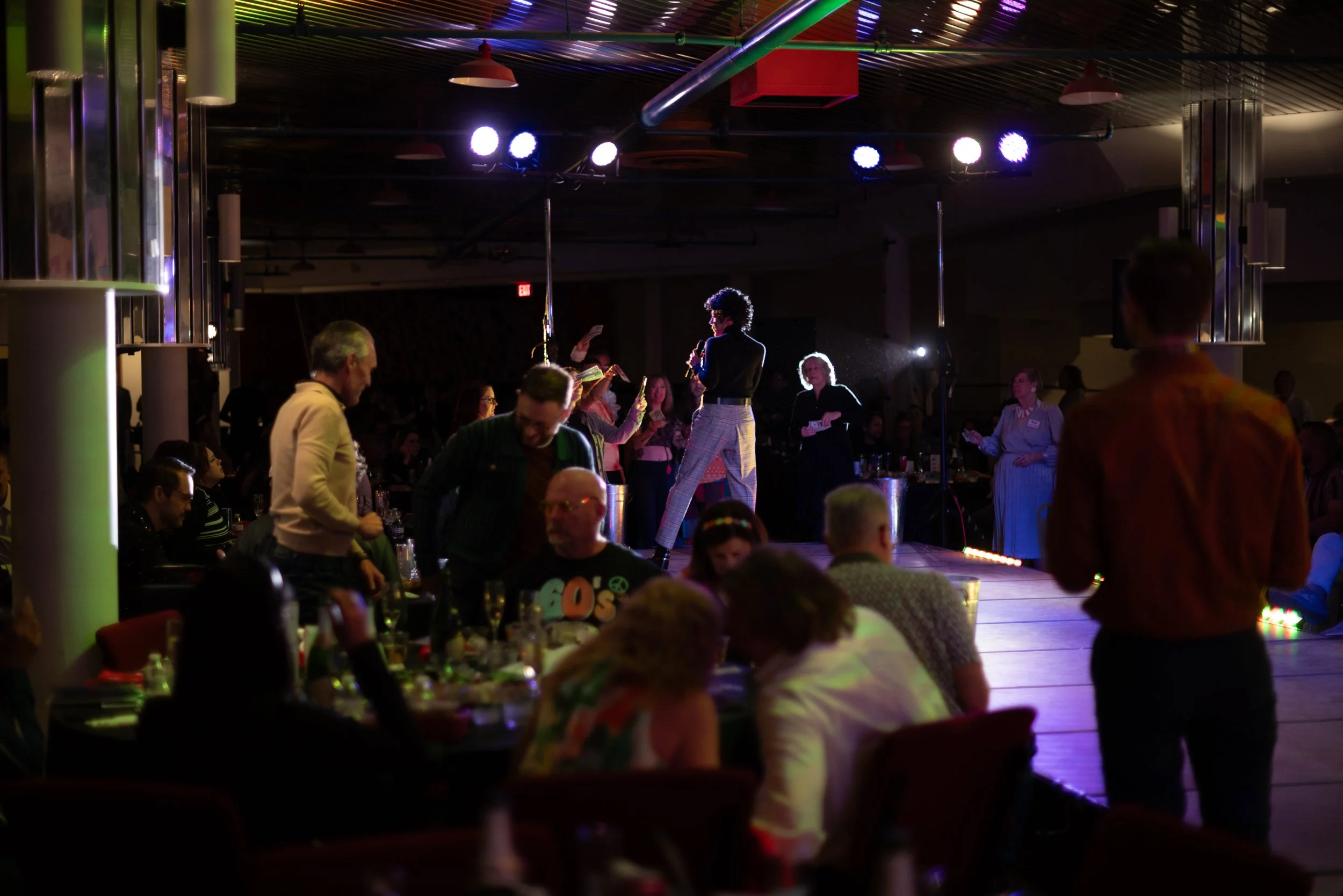 A woman standing on stage with a microphone, performing in a dimly lit event space with colorful lights, while audience members sit at tables and watch.