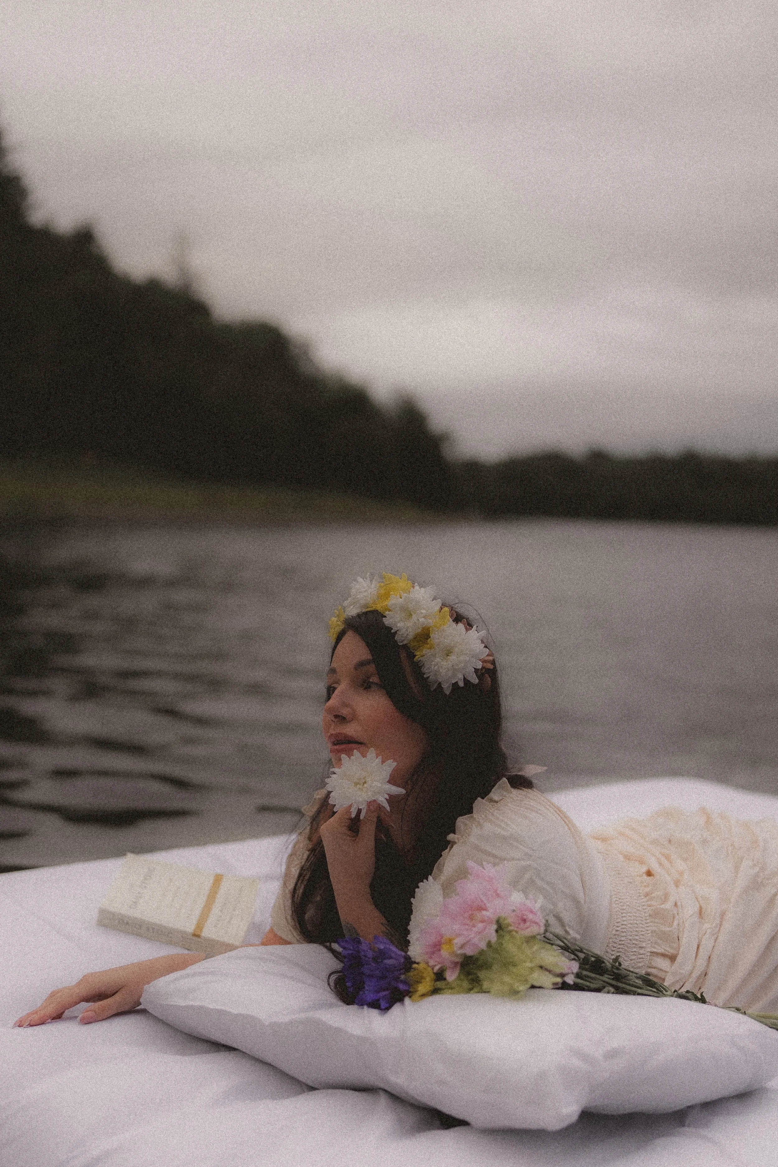 Woman lying on a white surface by a body of water with a forested hill in the distance, wearing a floral headband and holding flowers.